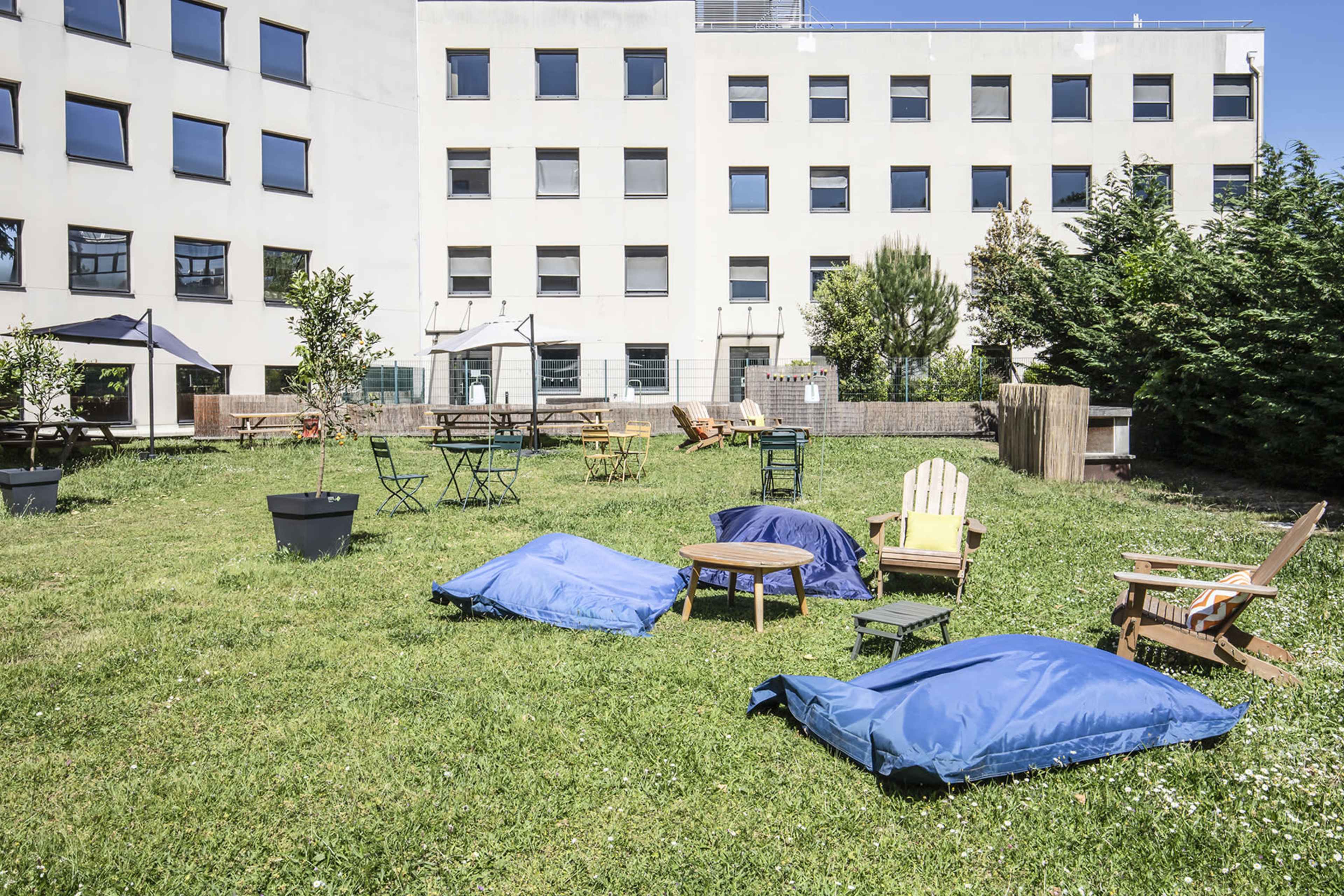 A grassy outdoor area with various seating arrangements, including blue bean bags, wooden chairs, and tables, adjacent to a white building.