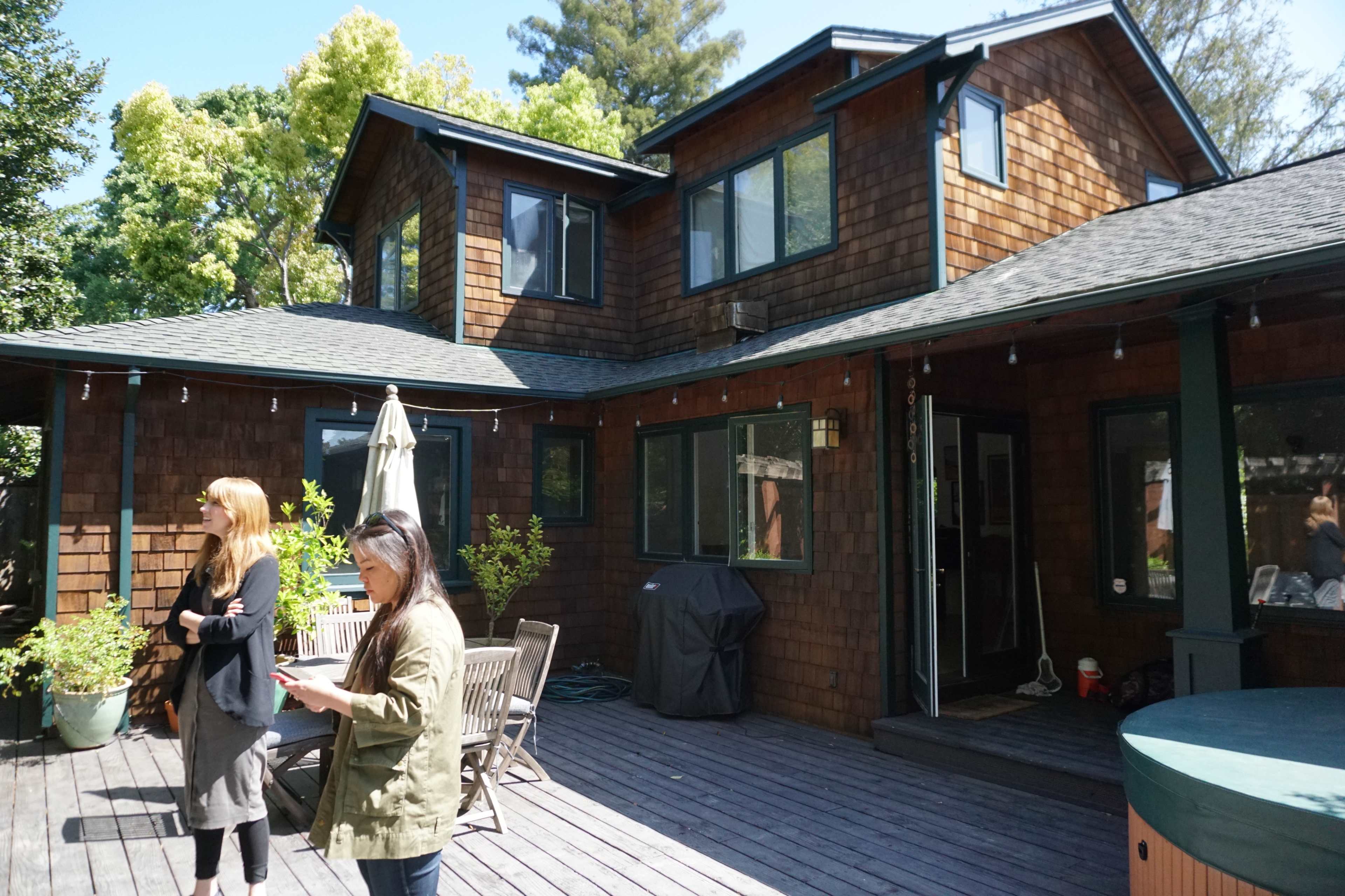 Two women stand on a wooden deck in front of a house featuring a multi-level, shingled exterior and large windows.