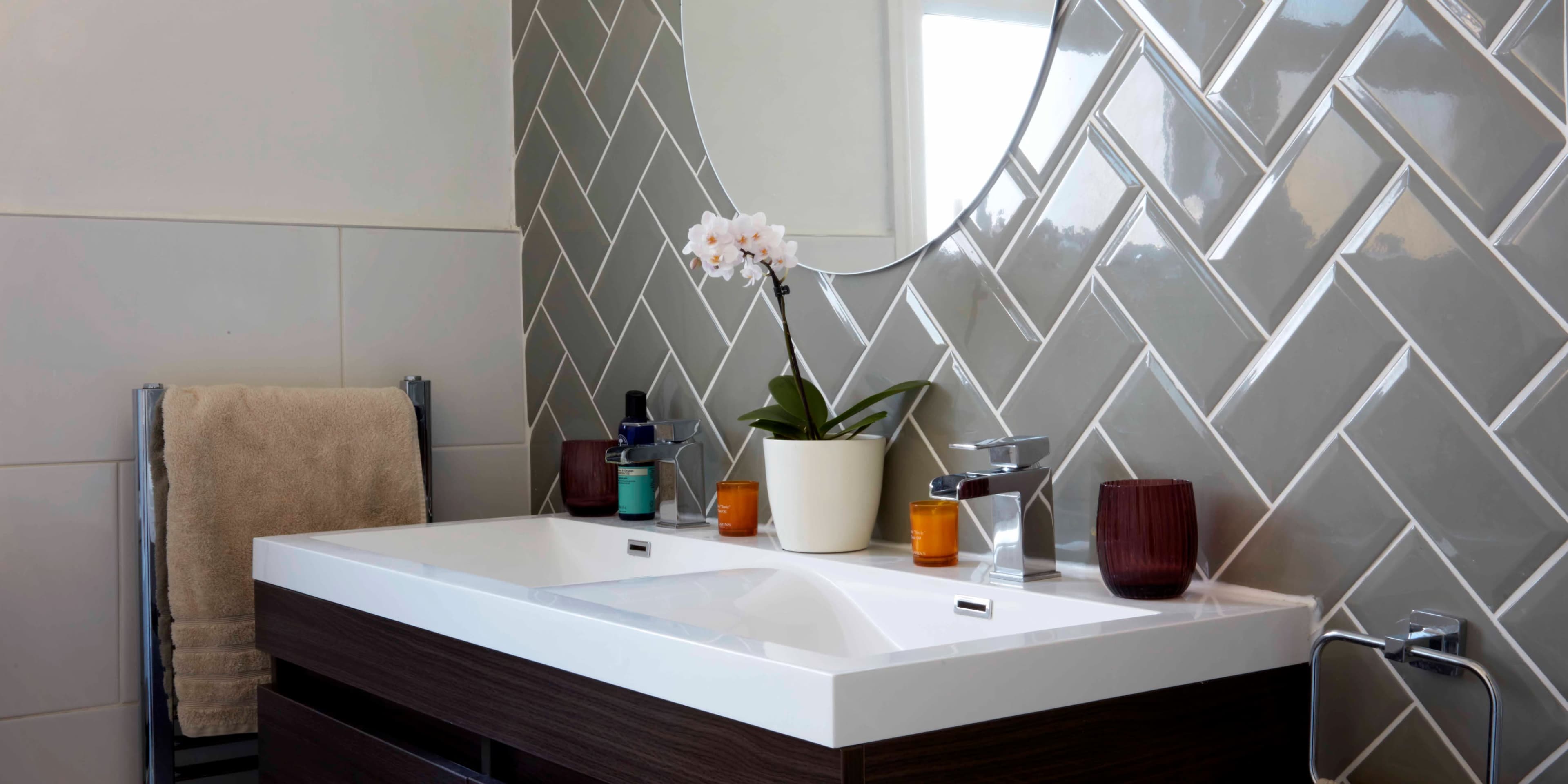 A modern bathroom features a sleek vanity with a white countertop, a round mirror, and decorative elements like an orchid and candles against a herringbone tile wall.