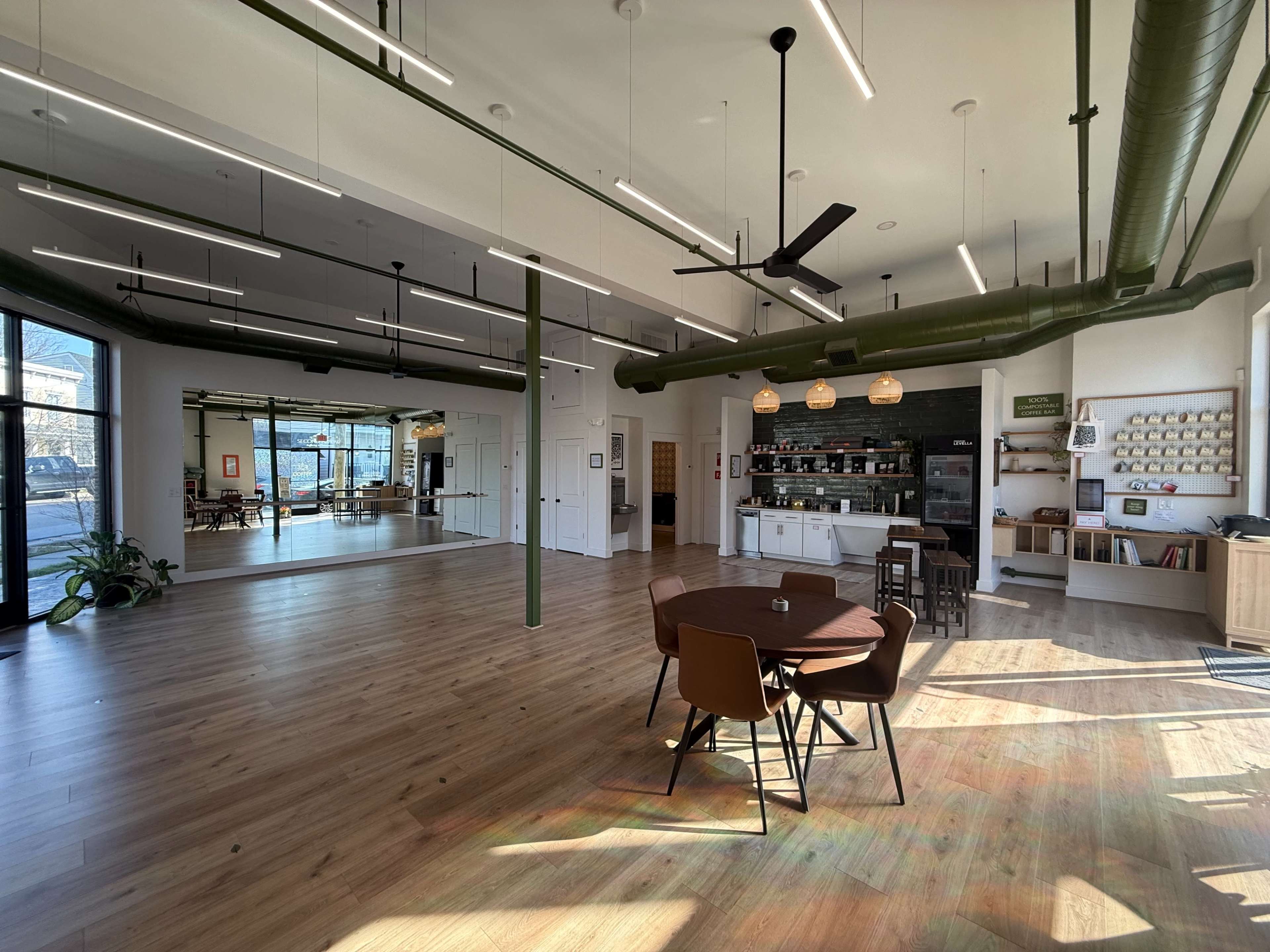 The image shows a spacious, well-lit interior of a modern open room featuring a dining table with chairs, a counter, shelves, and large windows.