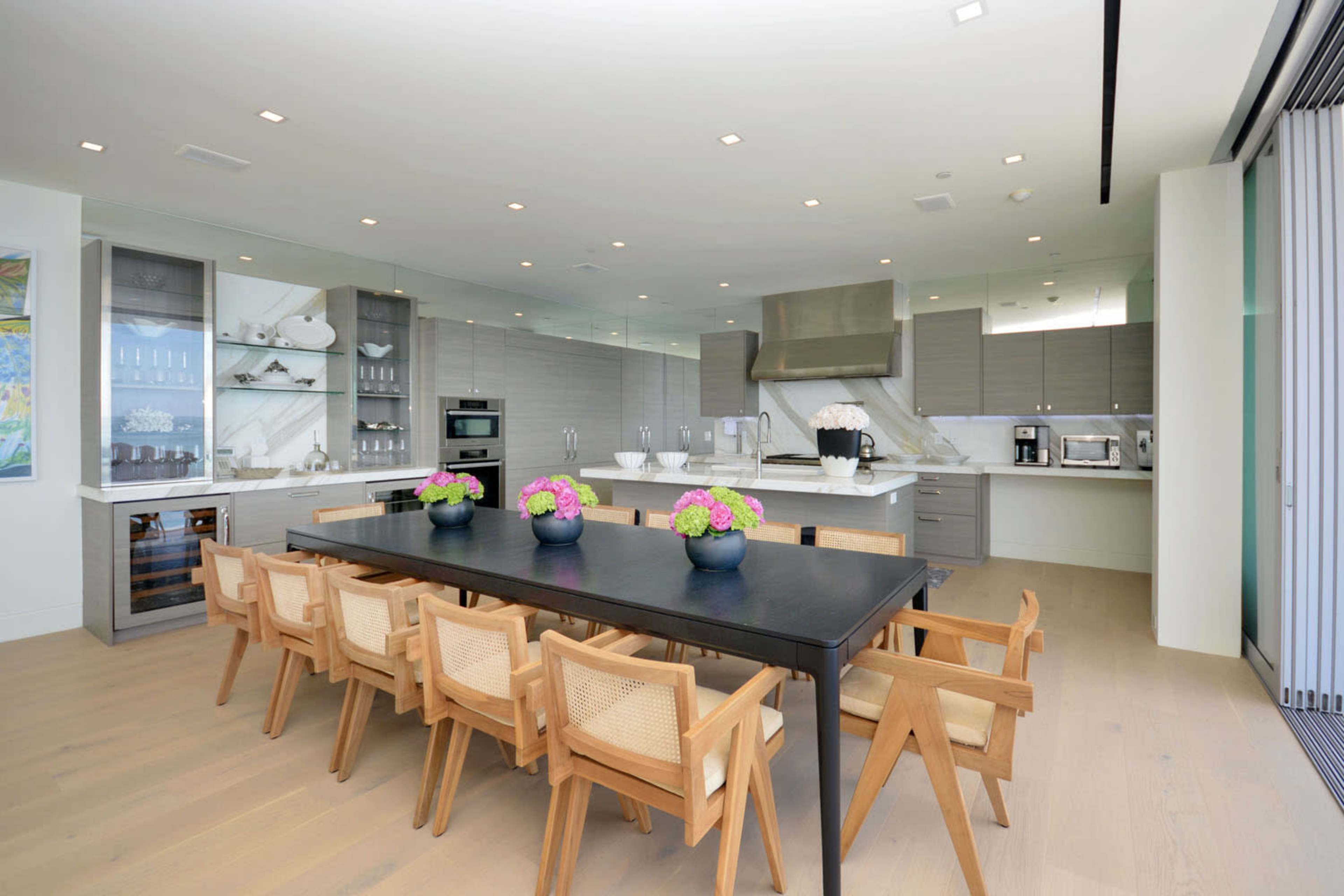 A modern kitchen and dining area features a large black table surrounded by wooden chairs, with a sleek kitchen equipped with stainless steel appliances and white cabinetry.