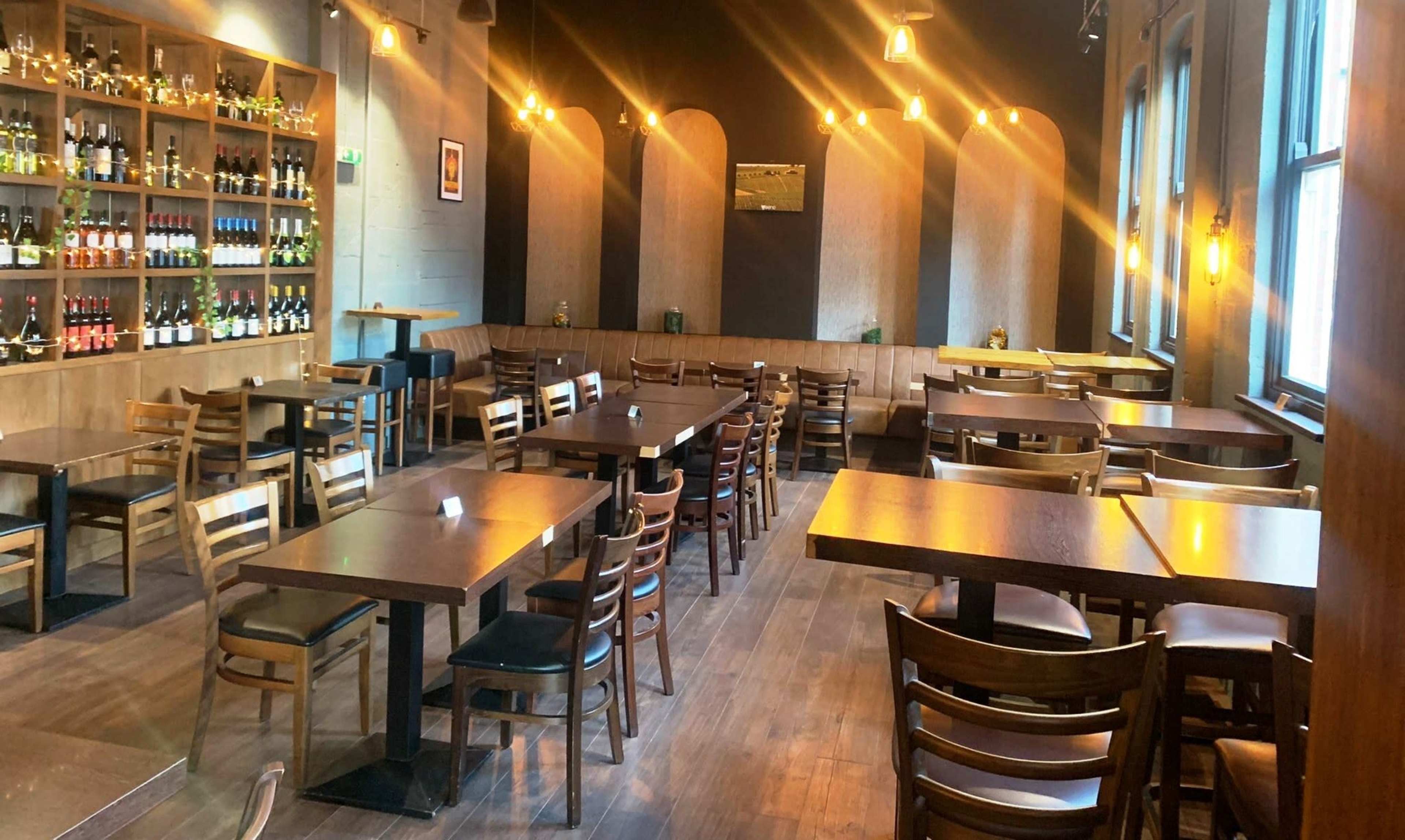 A spacious restaurant interior with wooden tables and chairs, shelves displaying bottles, and warm lighting.
