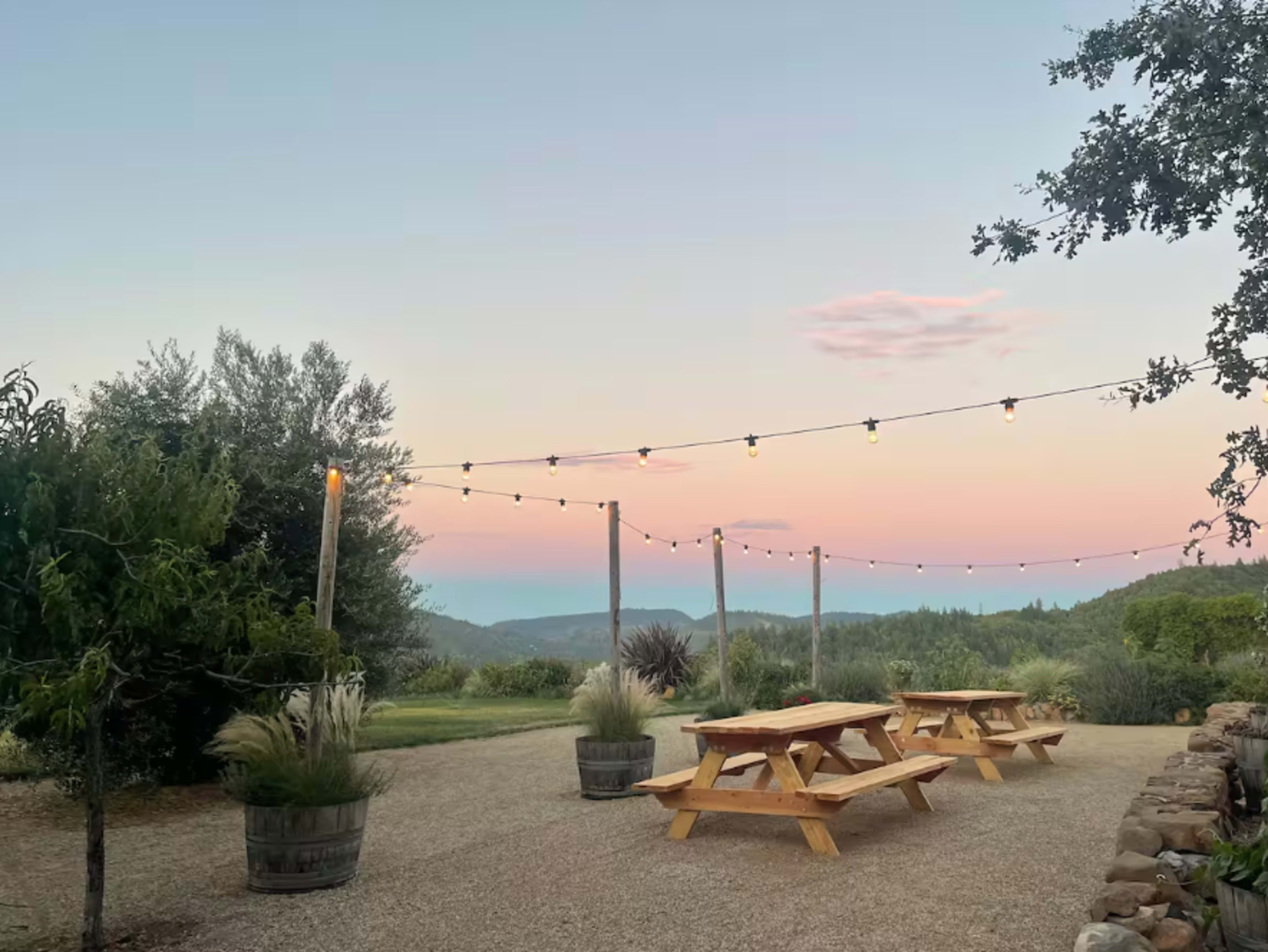 The image shows a picnic area with wooden tables and string lights set against a backdrop of rolling hills at sunset.
