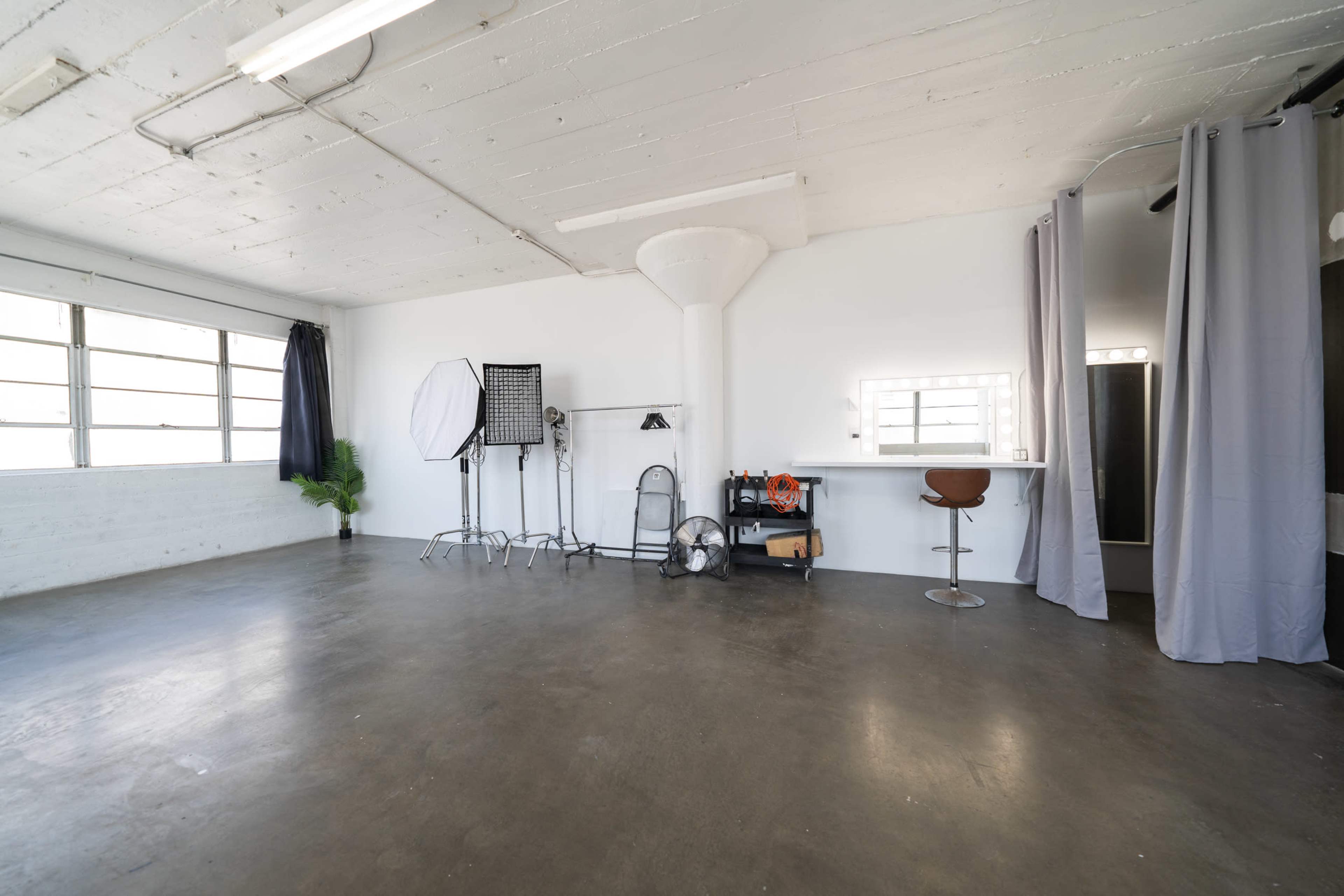 The image shows an empty studio space with a concrete floor, large windows, and lighting equipment arranged along one wall.