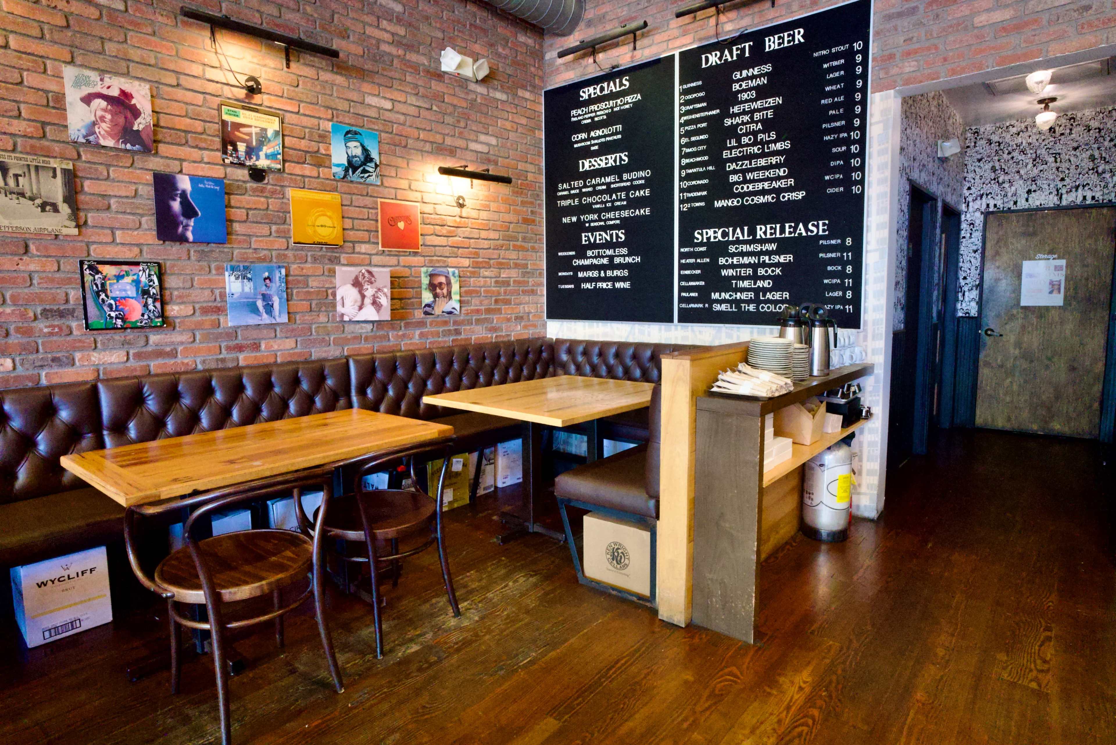 The scene depicts a cozy restaurant or café with wooden tables, a brick wall adorned with vintage album covers, and a large menu board listing specials and beverages.