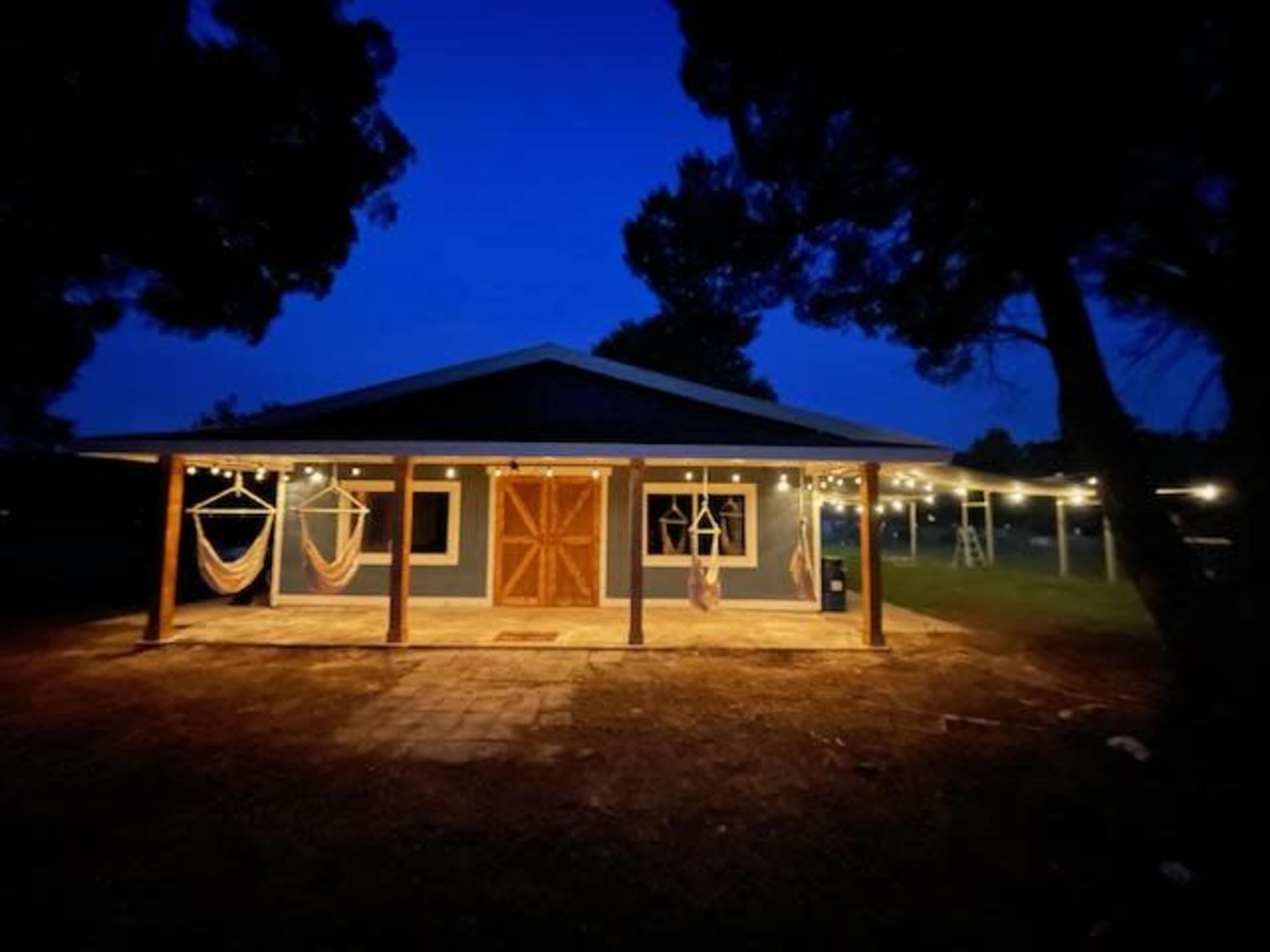 A well-lit house with a wooden front door and string lights features hammocks hanging on a porch at twilight.