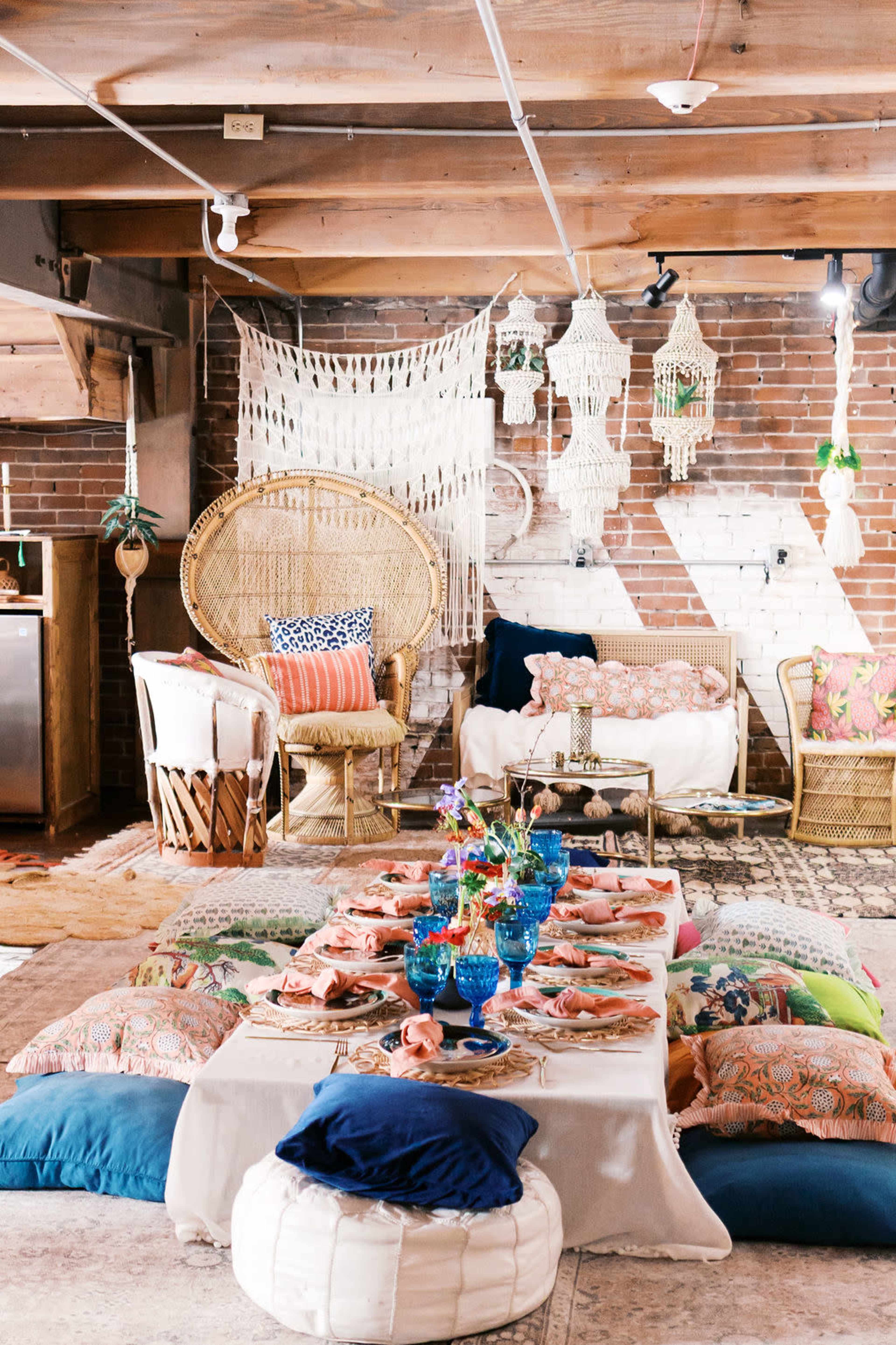 A cozy indoor space features a large, low table set with colorful plates and glasses, surrounded by cushions and decorative chairs.