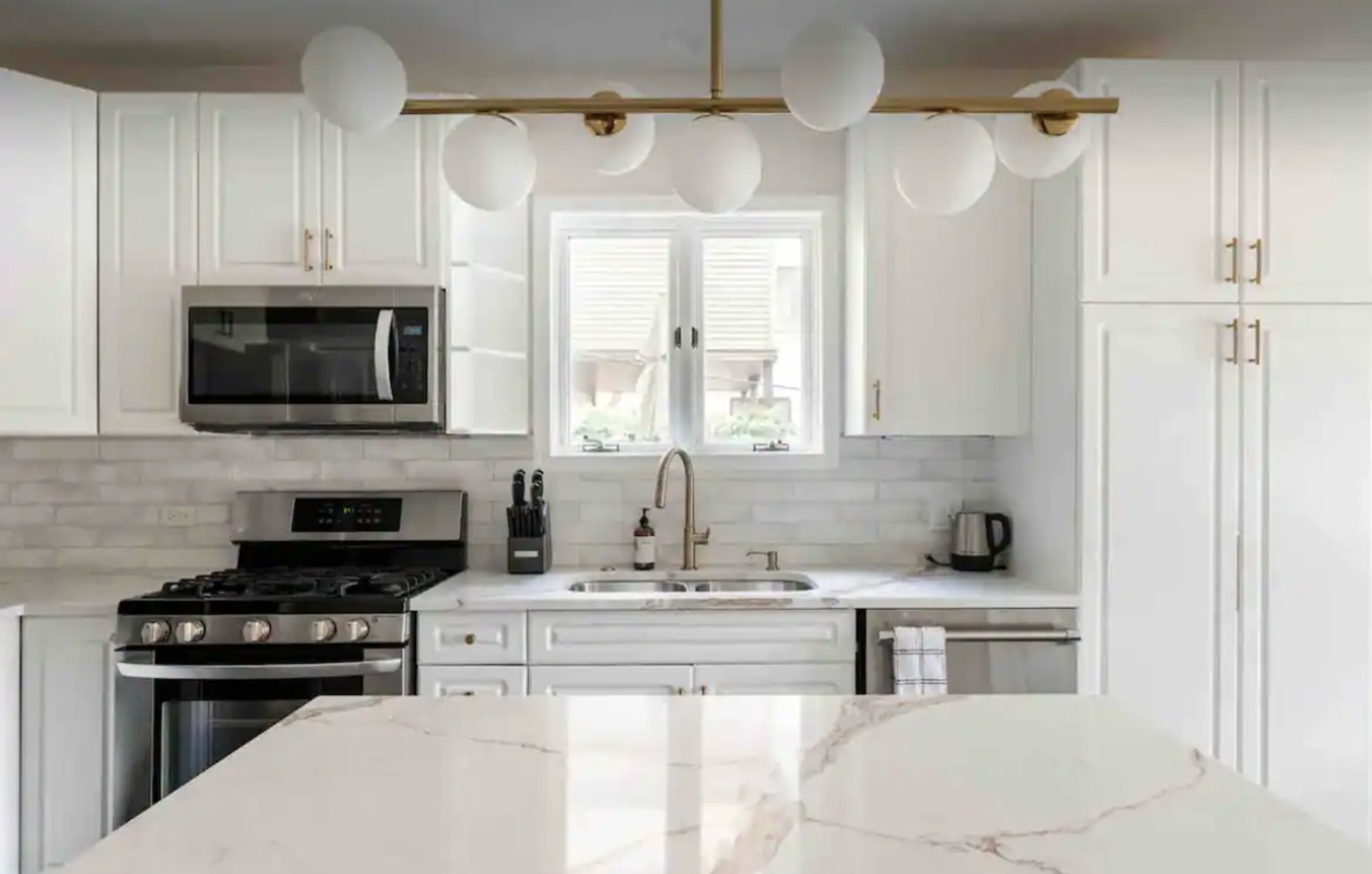 A modern kitchen features white cabinets, a marble countertop, stainless steel appliances, and a light fixture with spherical shades.