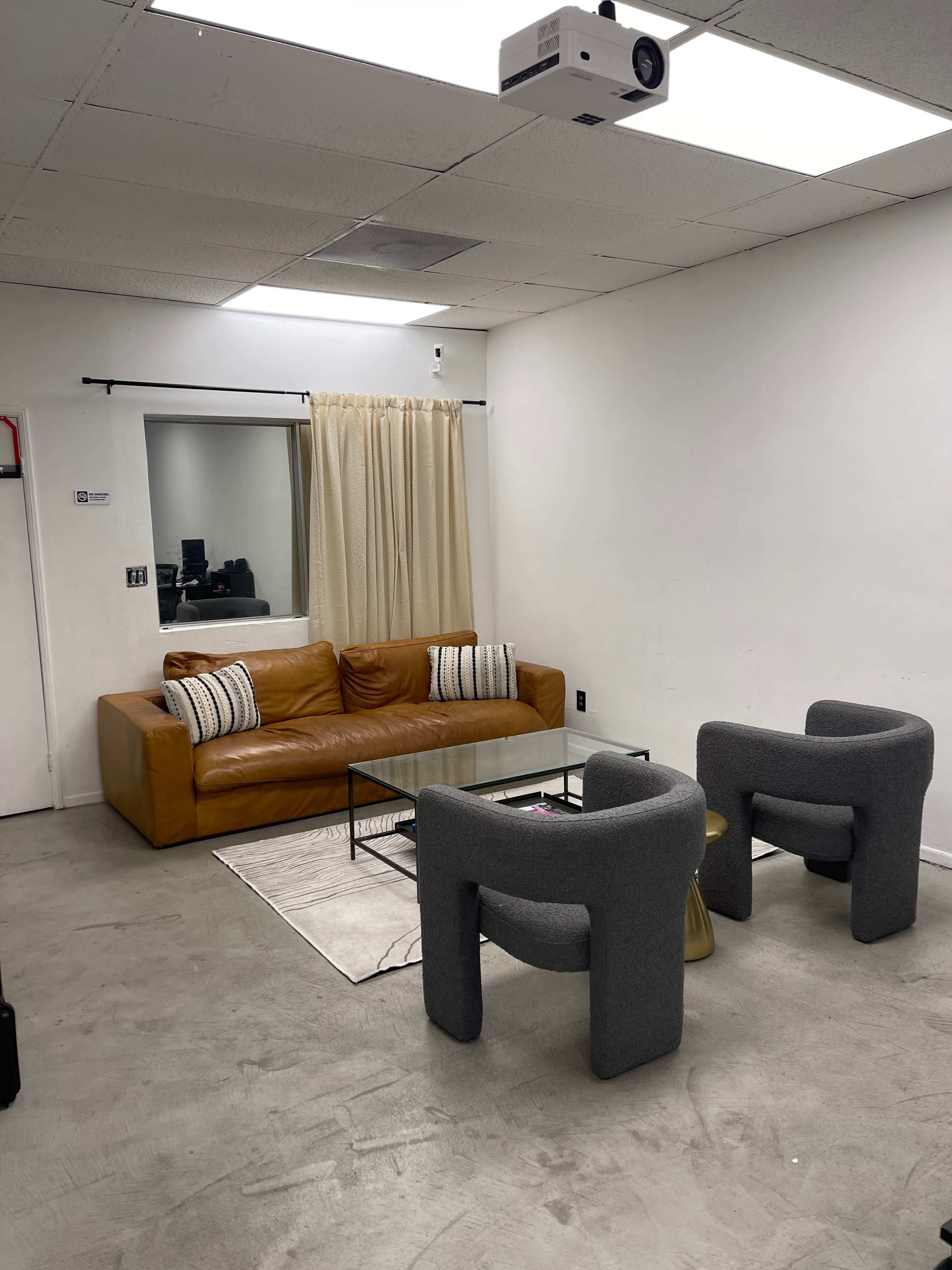 A simple room with a brown leather sofa, two gray armchairs, a glass coffee table, and a curtain covering a window.
