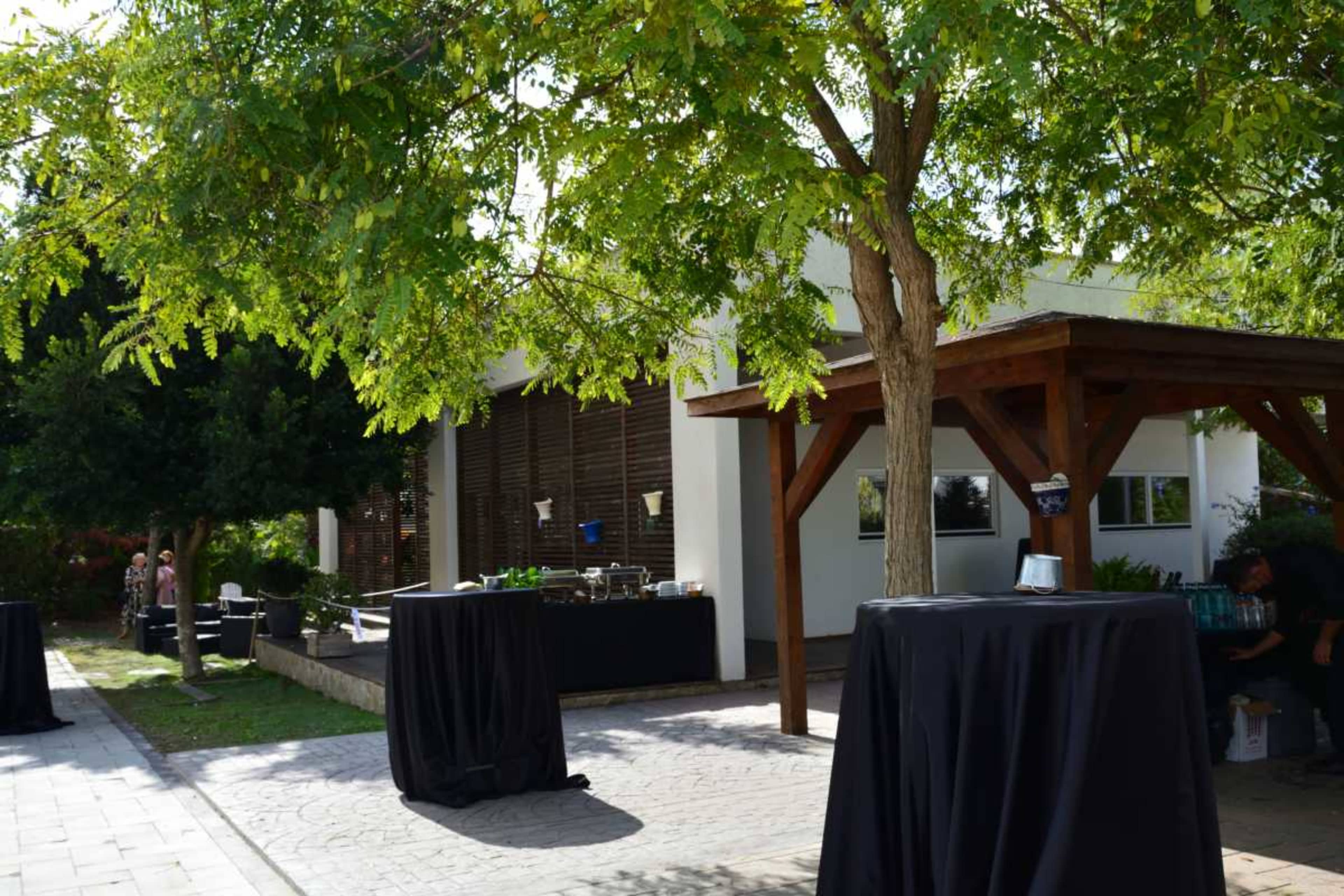The image shows an outdoor event area with tables covered in black cloth, surrounded by greenery and a building in the background.