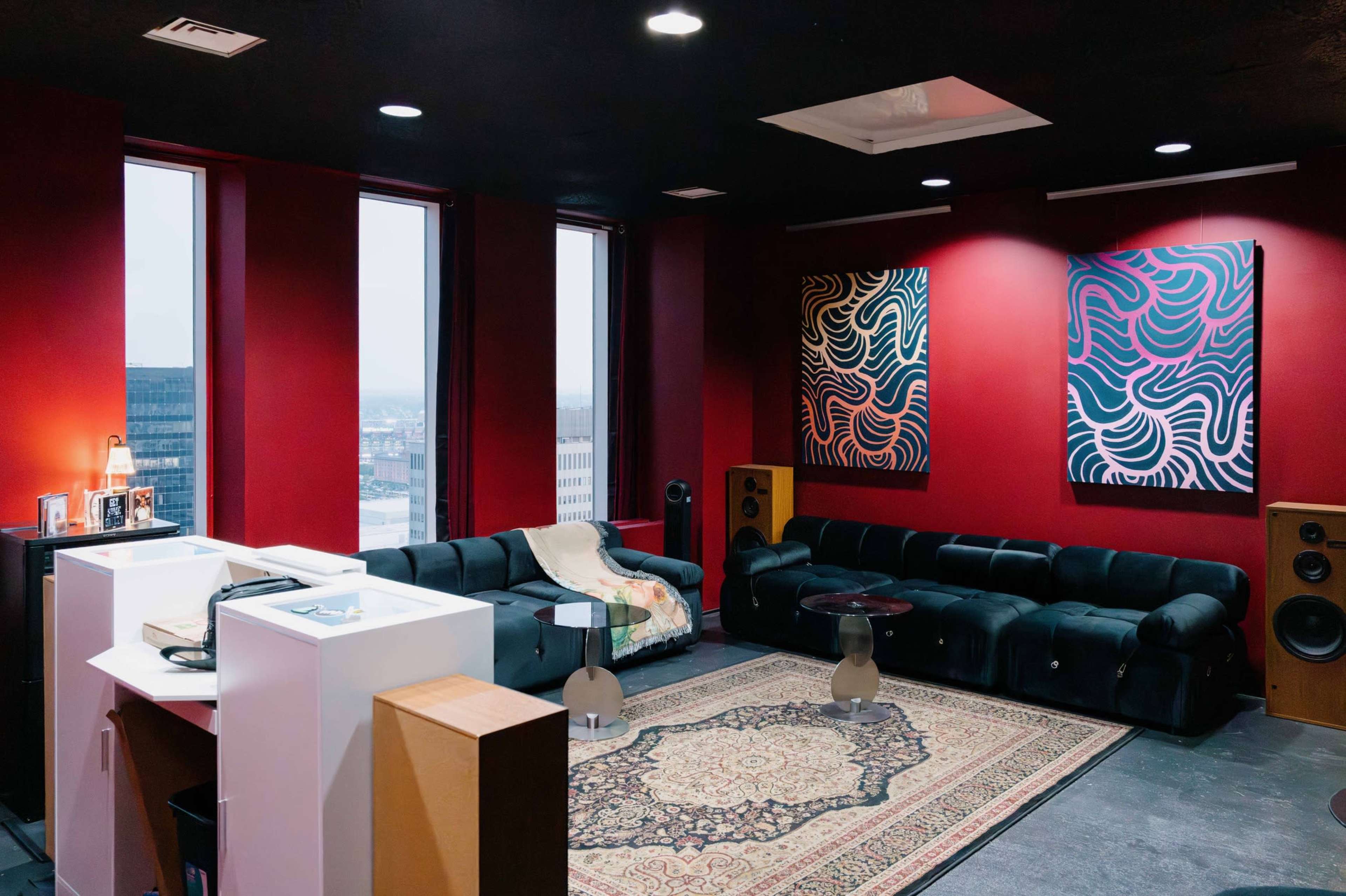 The image shows a modern lounge area with black sofas, two abstract paintings on the red walls, and a vintage rug on the floor.