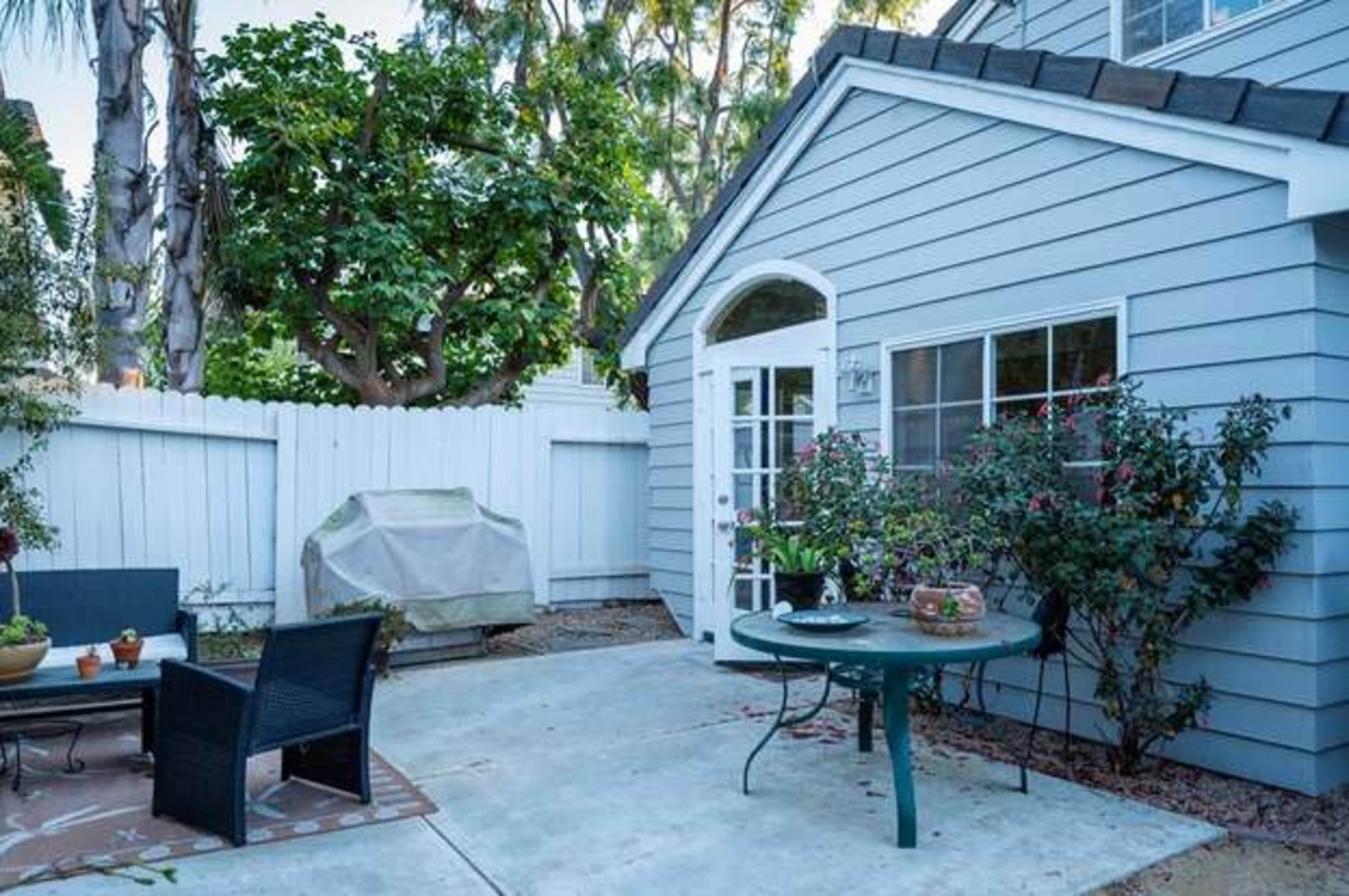A small backyard patio with a table and chairs, surrounded by a white fence and greenery.