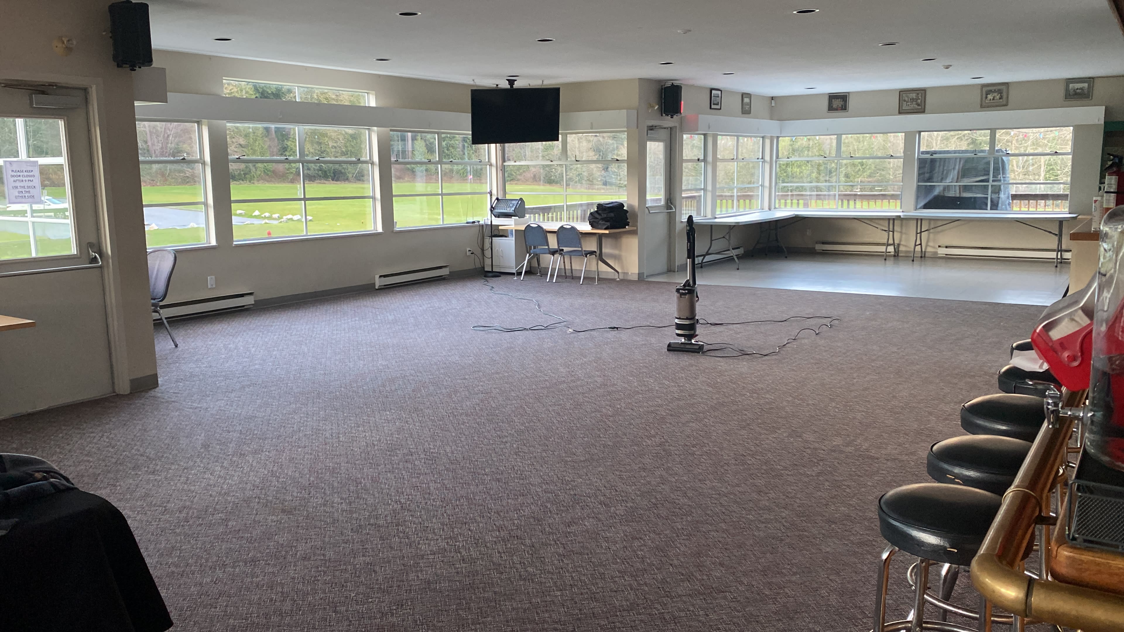 The image shows an empty room with large windows, a flat-screen TV mounted on the wall, and a vacuum cleaner on the floor, set up for a gathering or event.