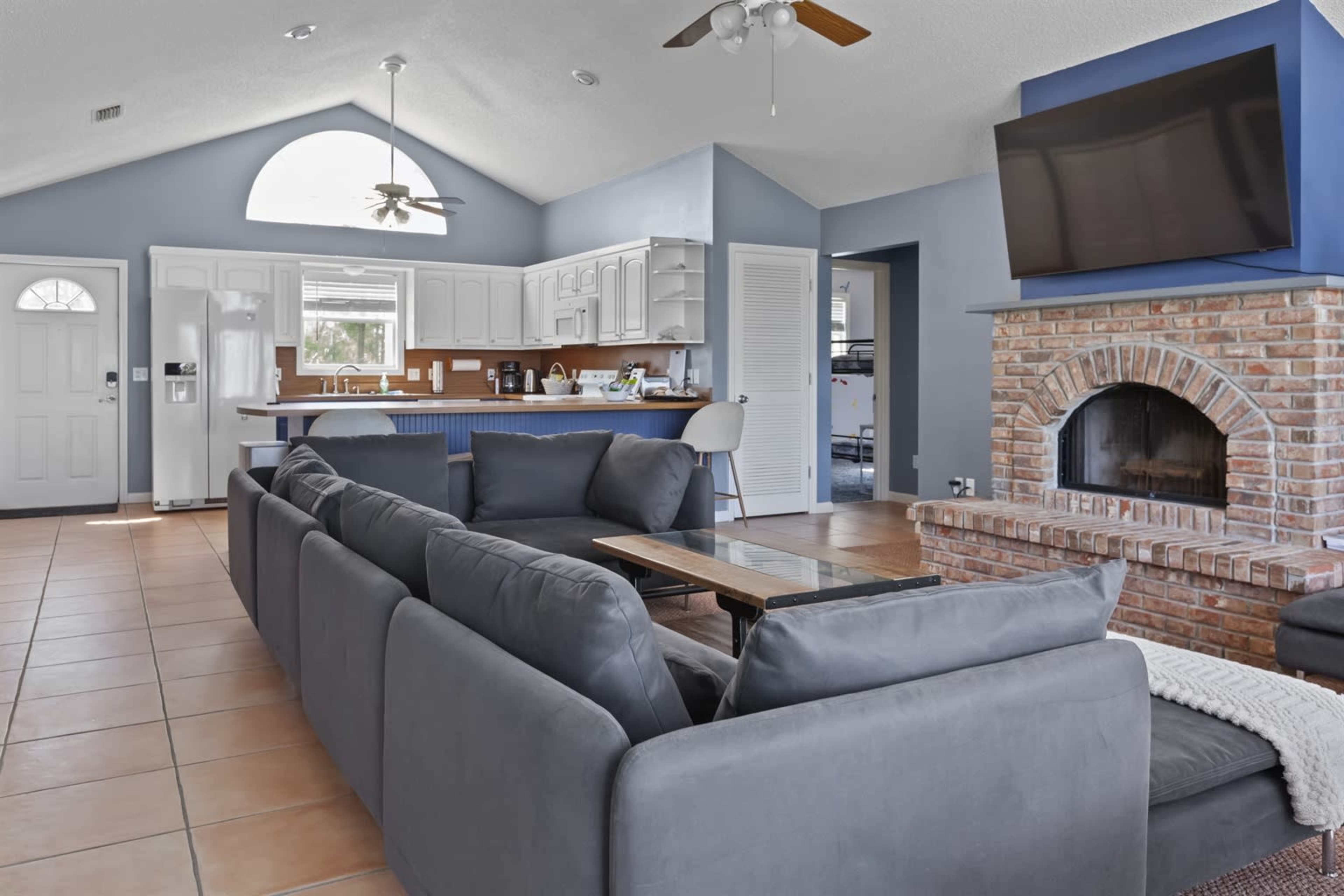 The image shows a living room with a sectional sofa, a brick fireplace, and a kitchen area visible in the background.