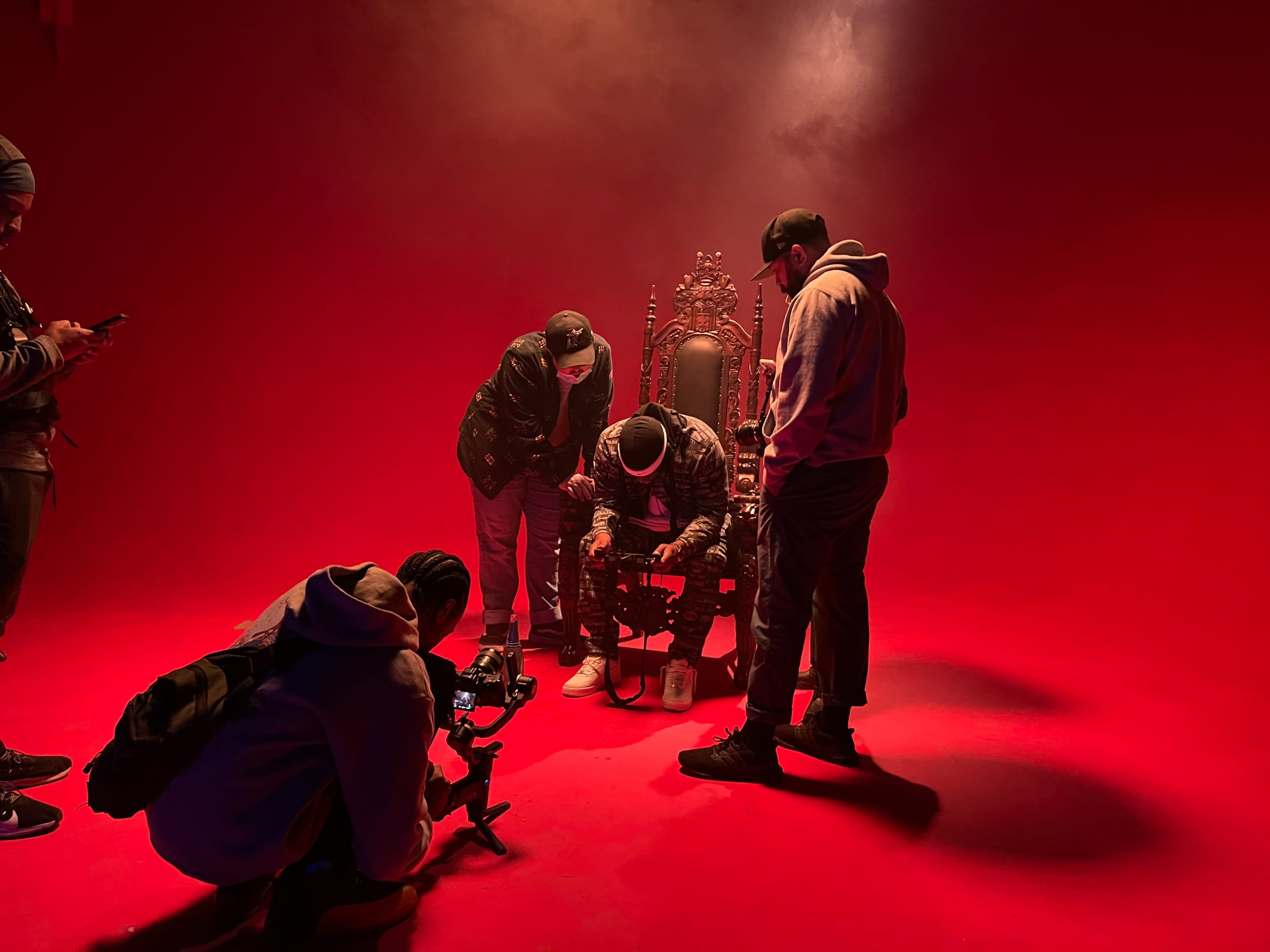 A group of people is gathered around an individual seated on an ornate throne against a red backdrop, with smoke effects in the air.