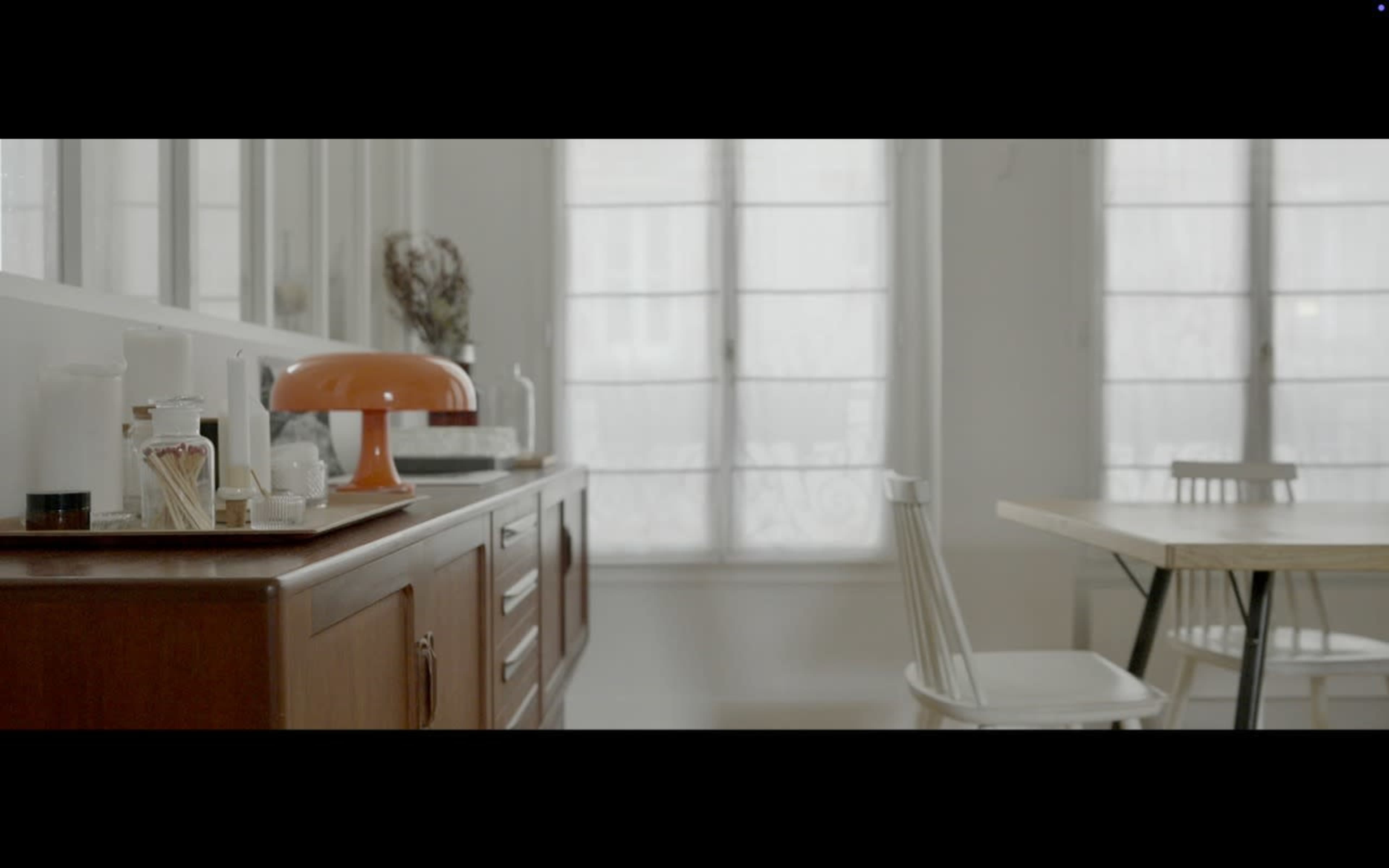 A wooden sideboard displays various containers and a lamp in a bright room with large windows and a dining table.
