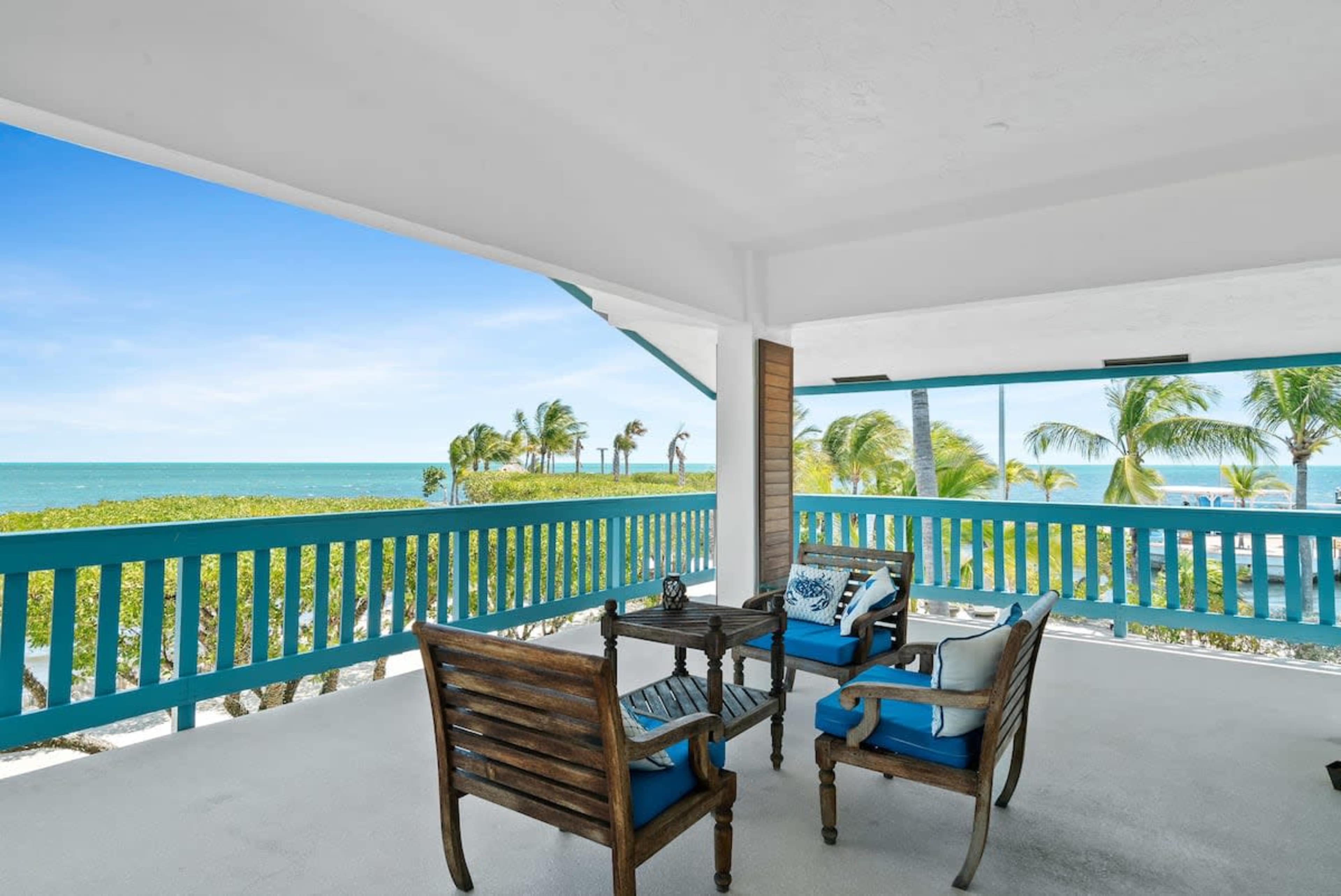 A spacious patio features wooden furniture and a clear view of the ocean and palm trees in the distance.