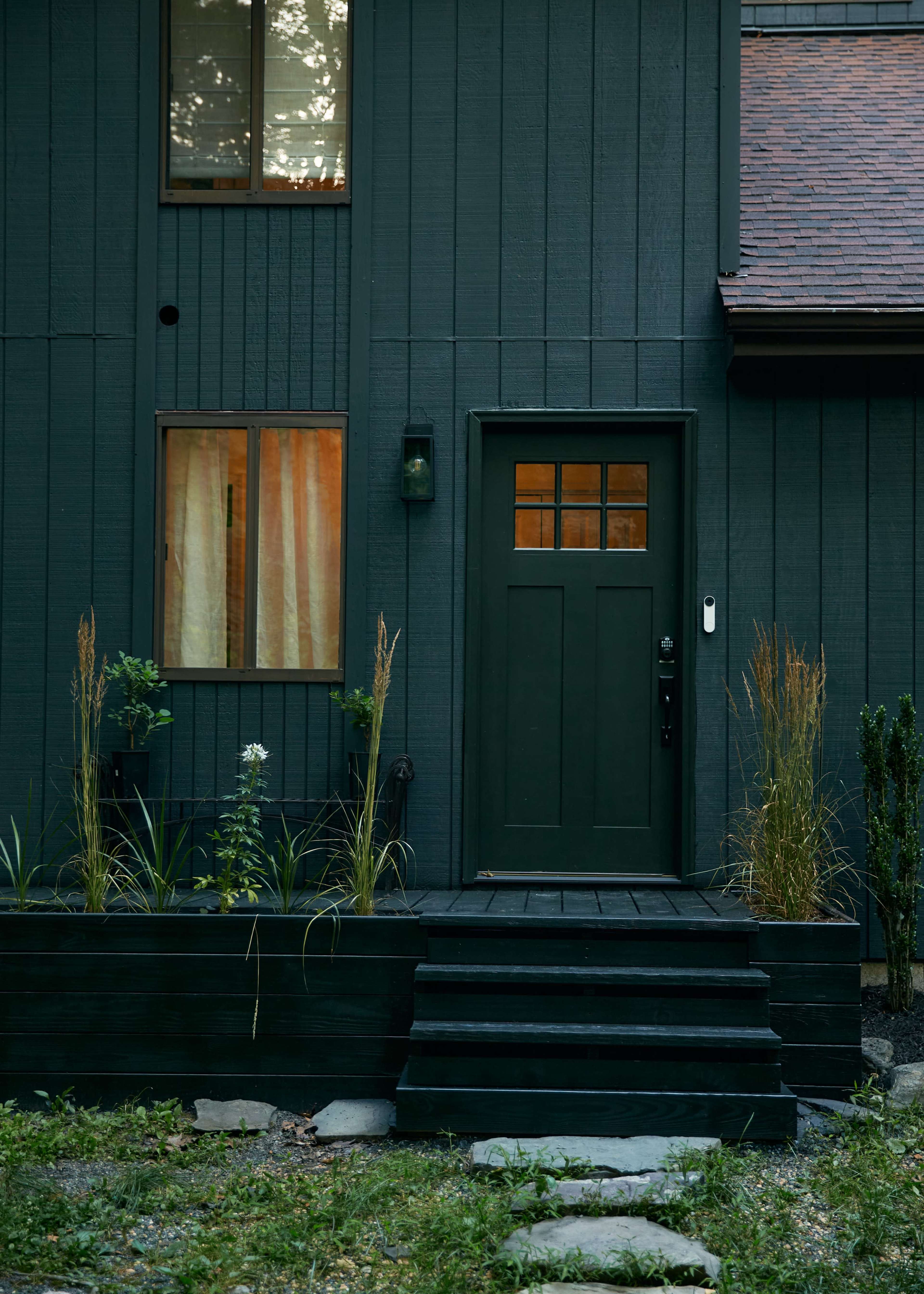 A dark wood-paneled house featuring a green front door, a window with sheer curtains, and a small landscaped entryway with plants and stones.