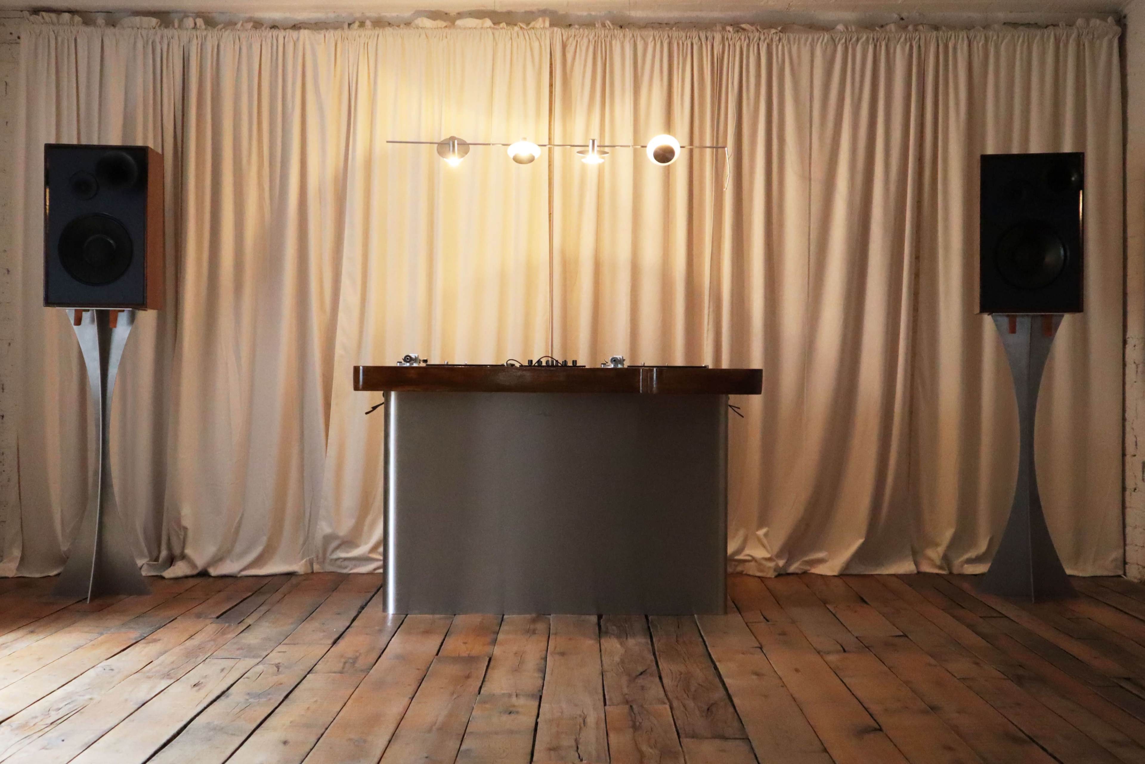 The image shows a minimalist DJ setup featuring a rectangular wooden table with a turntable in the center and two large speakers positioned on either side against a backdrop of draped fabric.