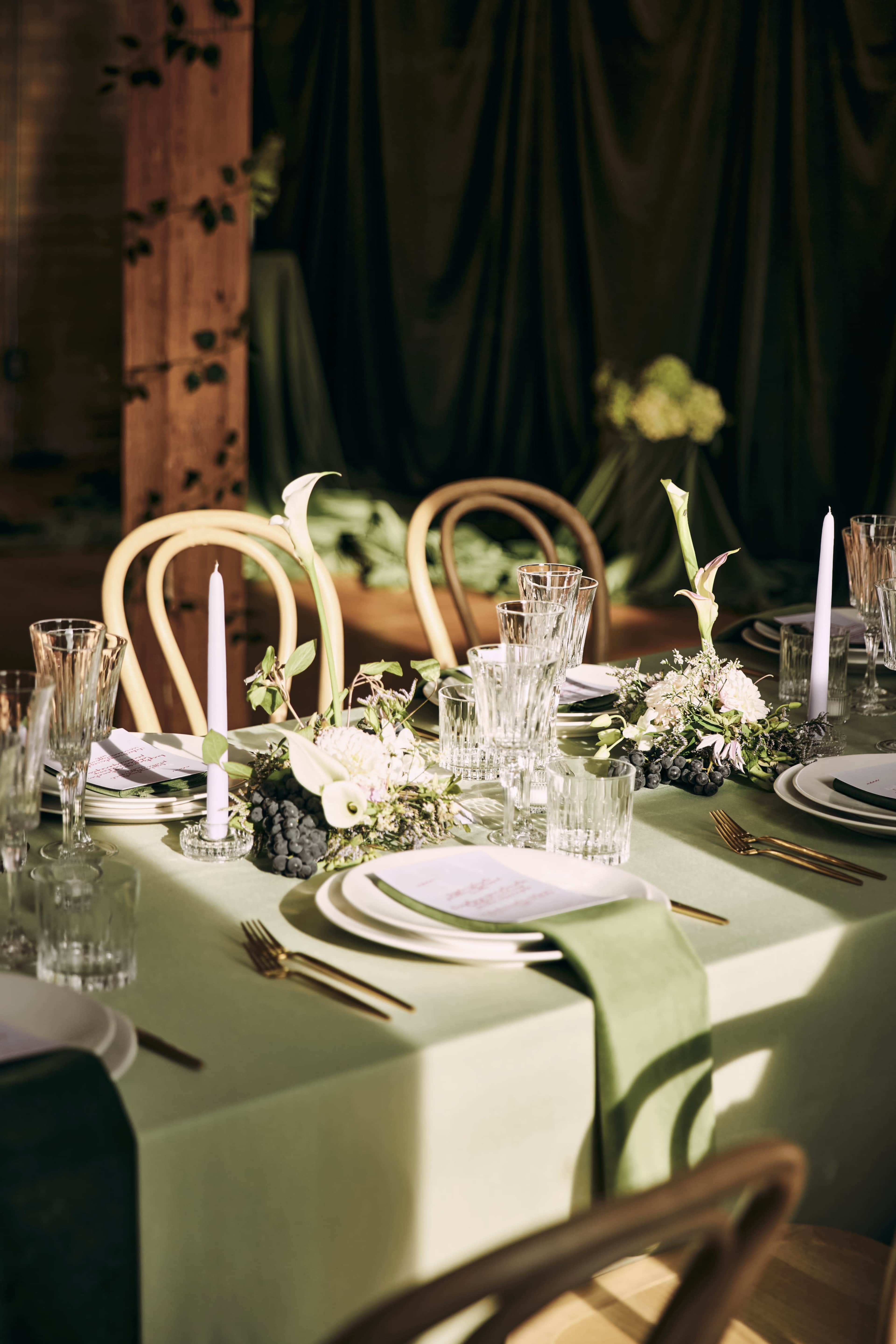 A neatly arranged dining table features a soft green tablecloth, elegant dishware, glassware, and floral decorations, illuminated by natural light.