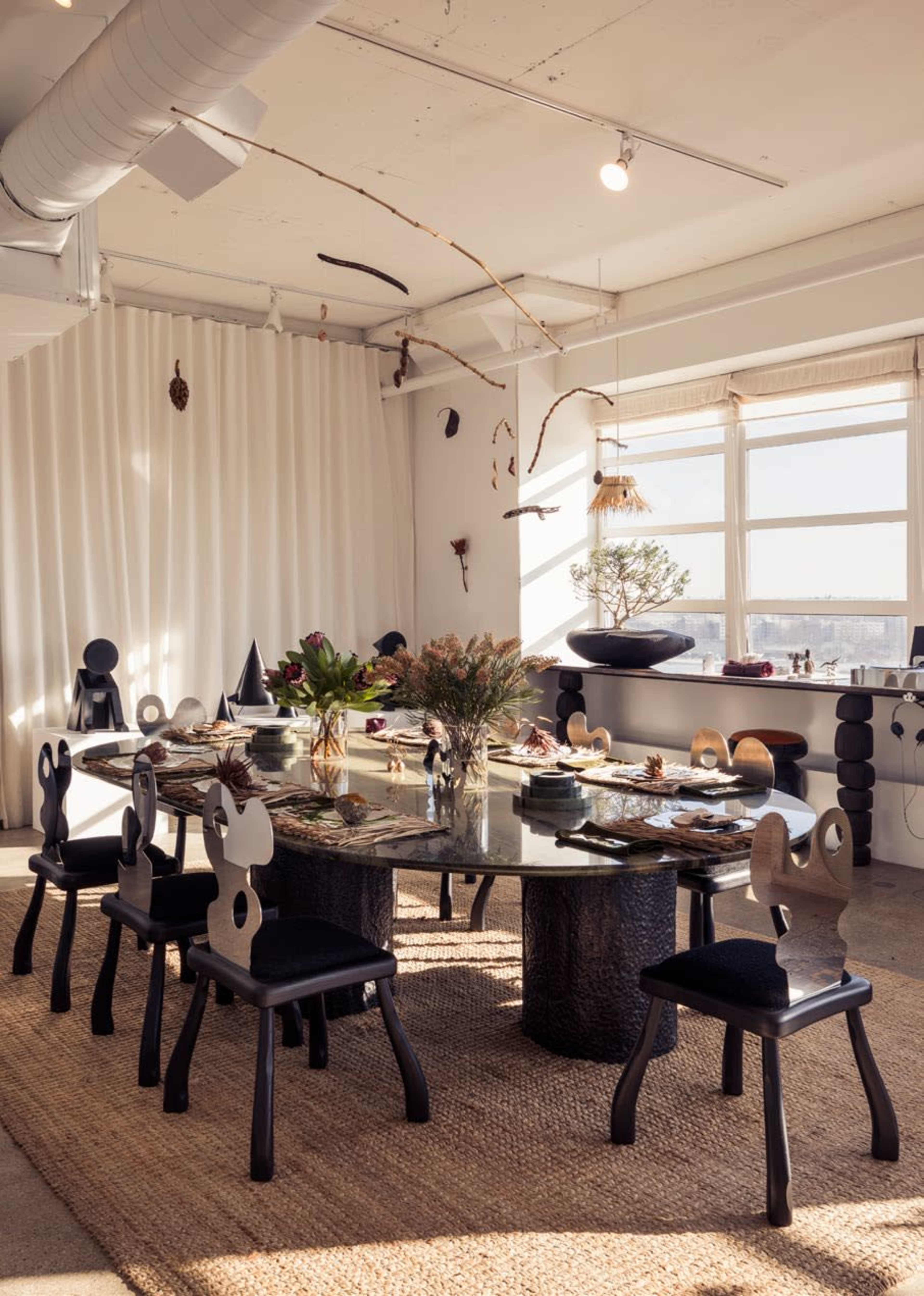 A large, round dining table is set with eight chairs in a bright room featuring large windows and decorative elements on the walls and ceiling.