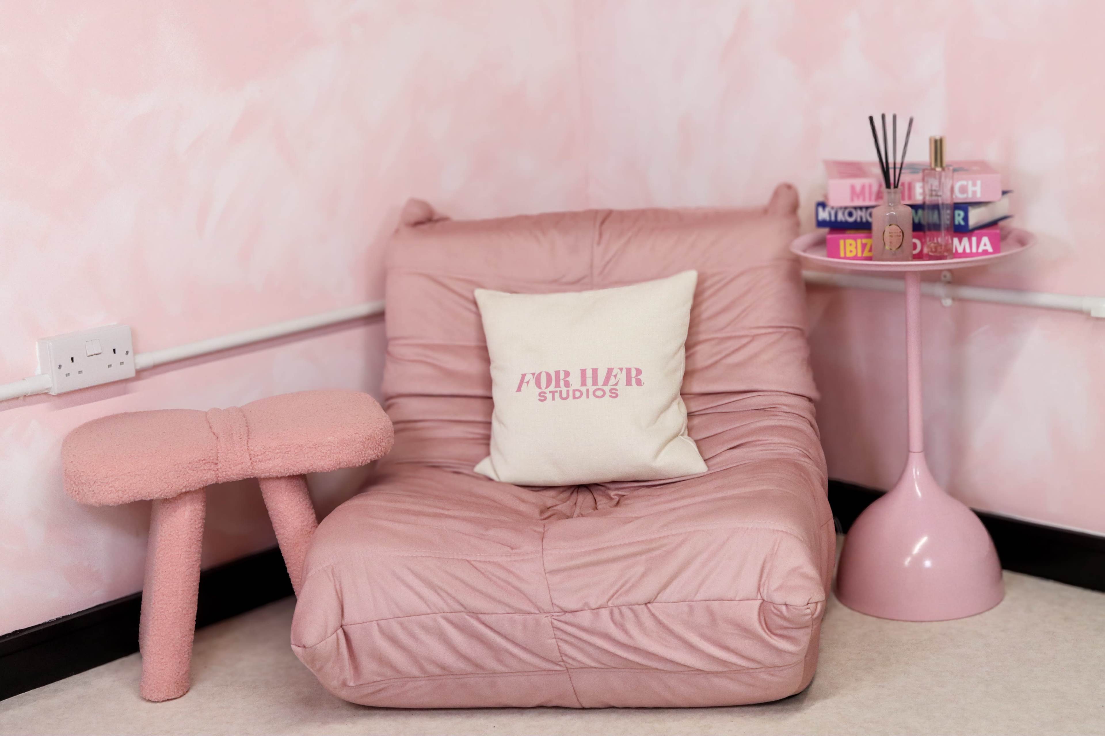 The image shows a pink-themed corner with a plush chair, a decorative cushion, and a small side table featuring books and a reed diffuser.