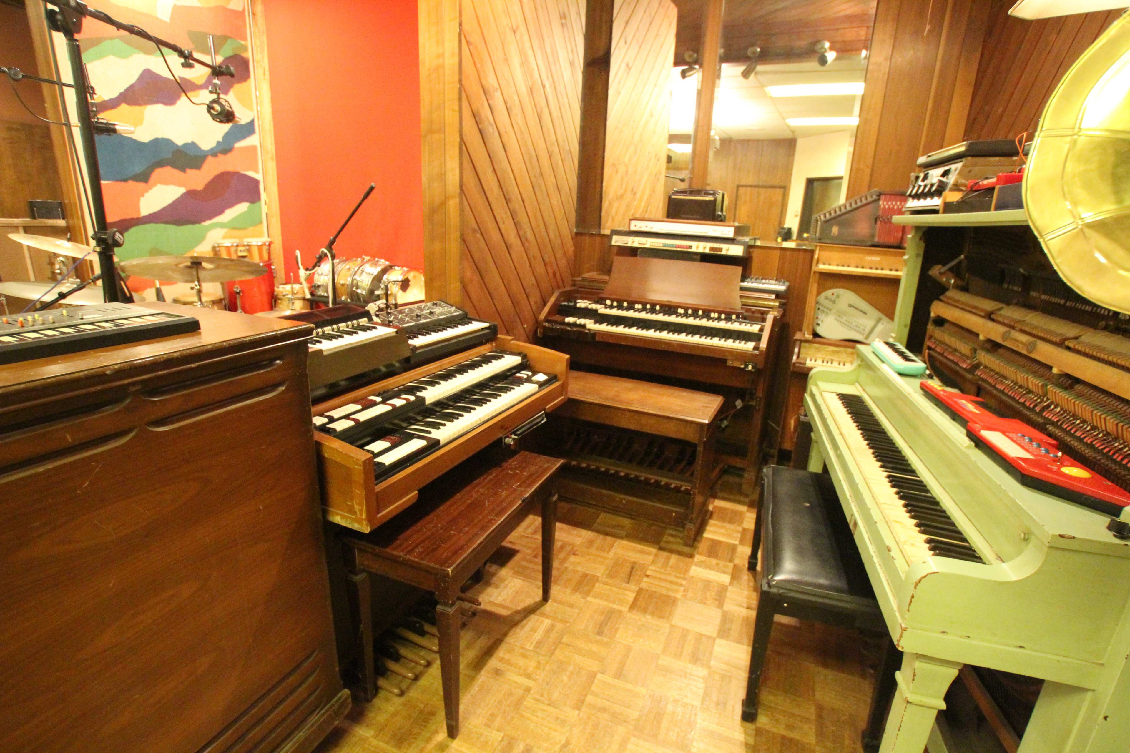 A room filled with various vintage keyboards and musical instruments, including electric organs and synthesizers, arranged around wooden furniture.