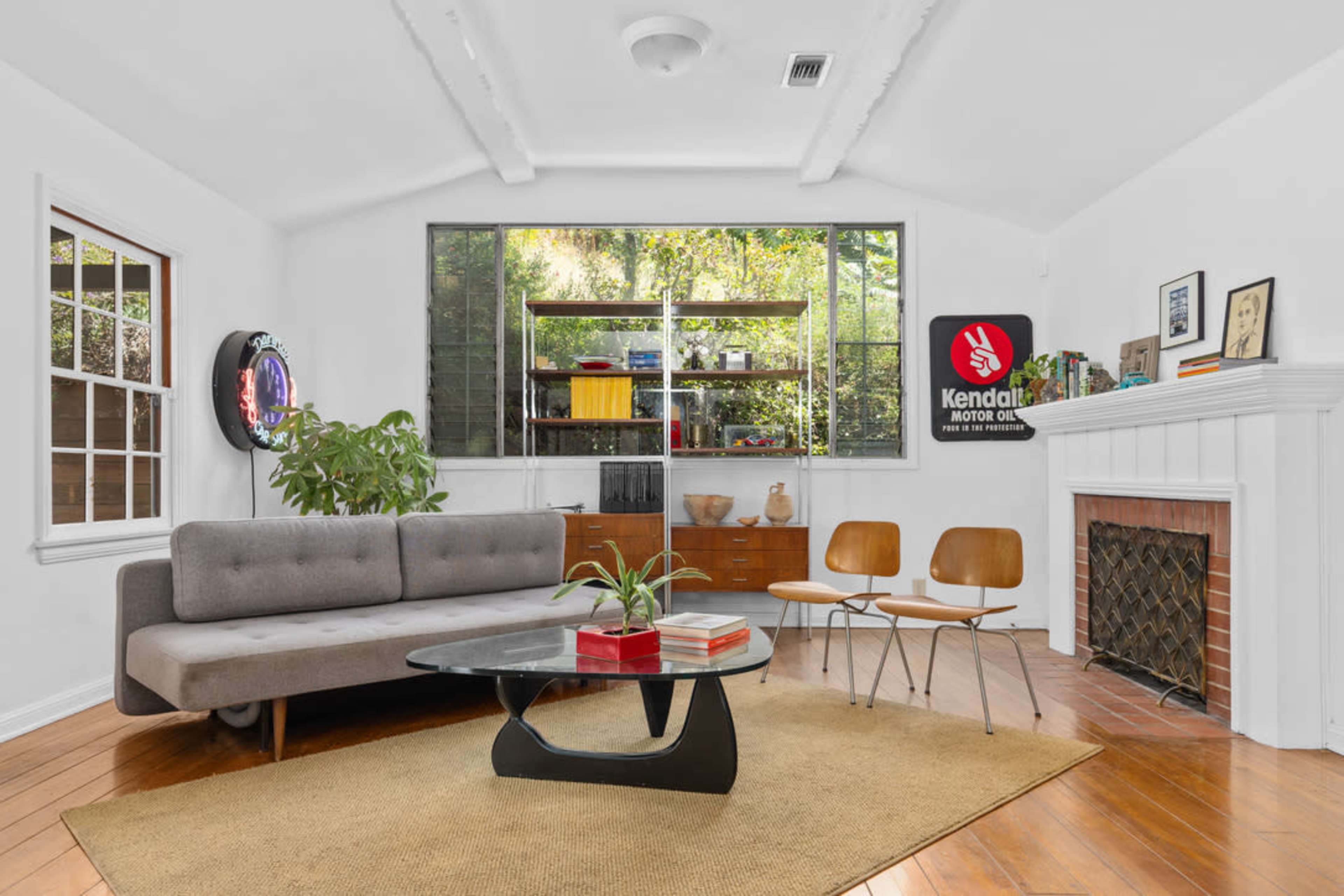 The image shows a bright living room featuring a gray sofa, wooden chairs, a glass coffee table, and shelves adorned with books and decorative items.