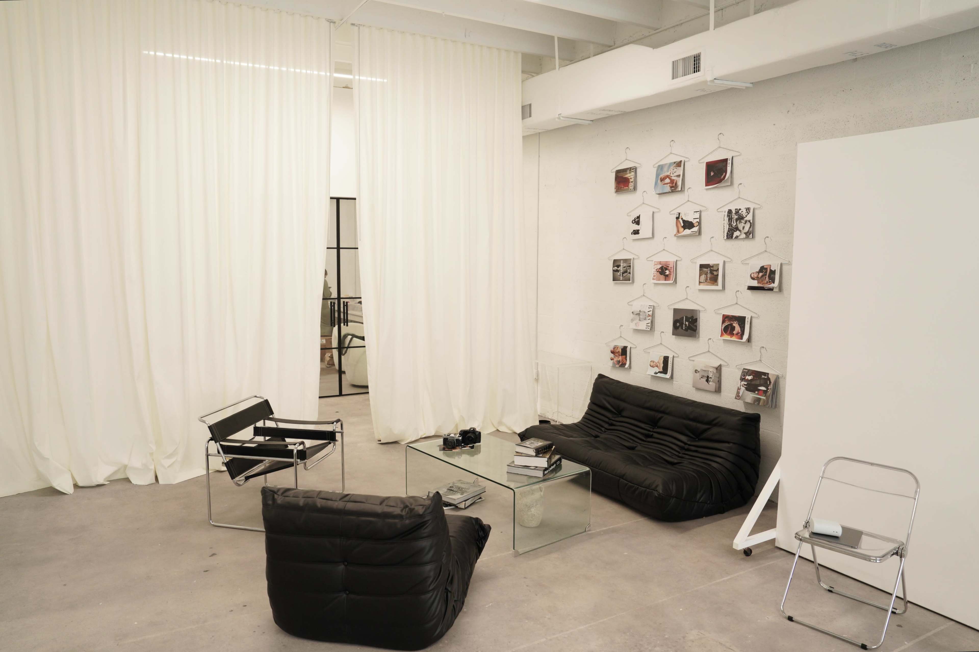 A minimalist room with two black chairs, a glass coffee table, sheer white curtains, and a wall displaying framed photographs.