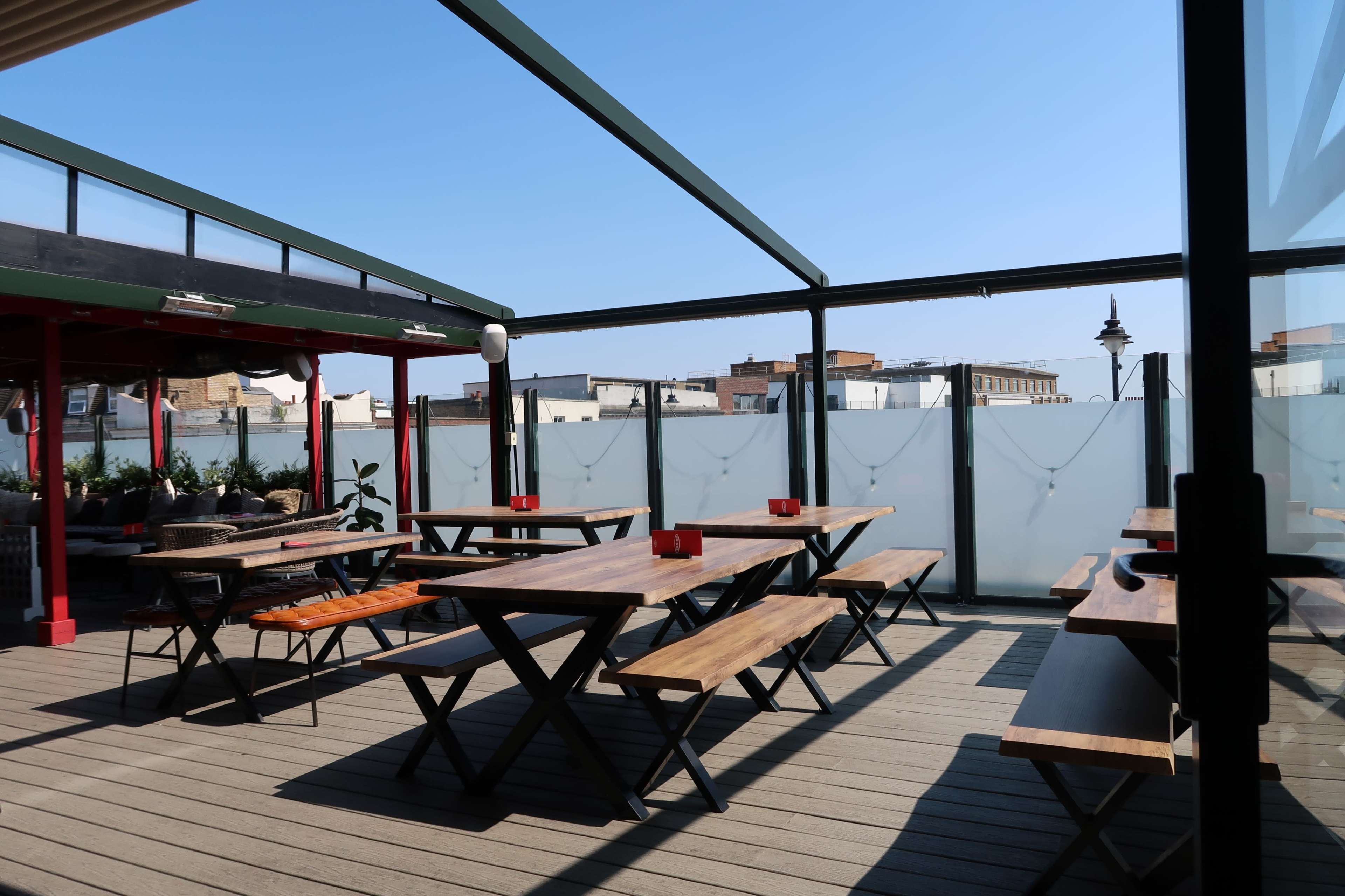 The image shows a rooftop terrace with wooden tables and benches under a clear blue sky.