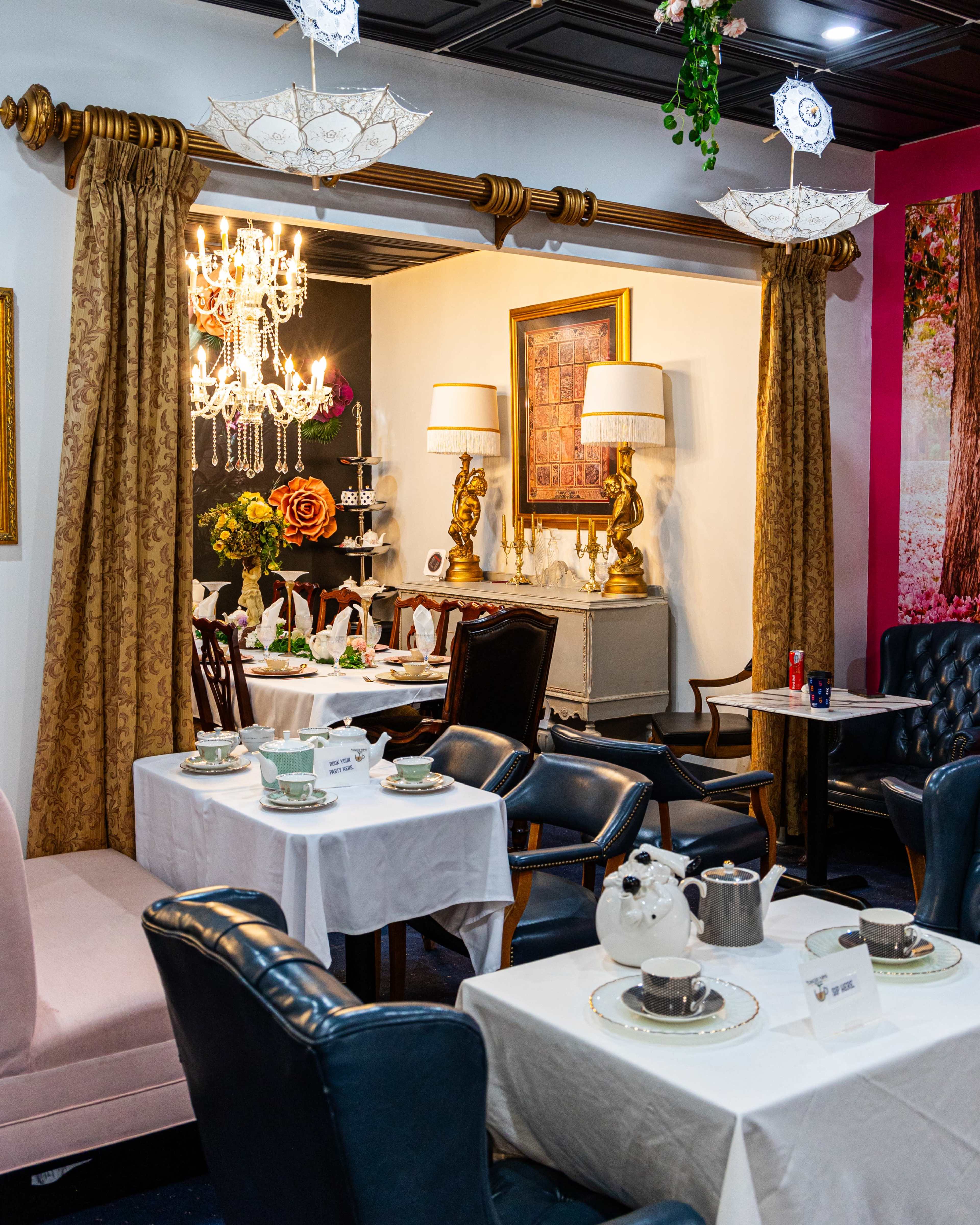 The image shows a cozy dining area with tables set for tea, surrounded by elegant decor including a chandelier, curtains, and decorative umbrellas.