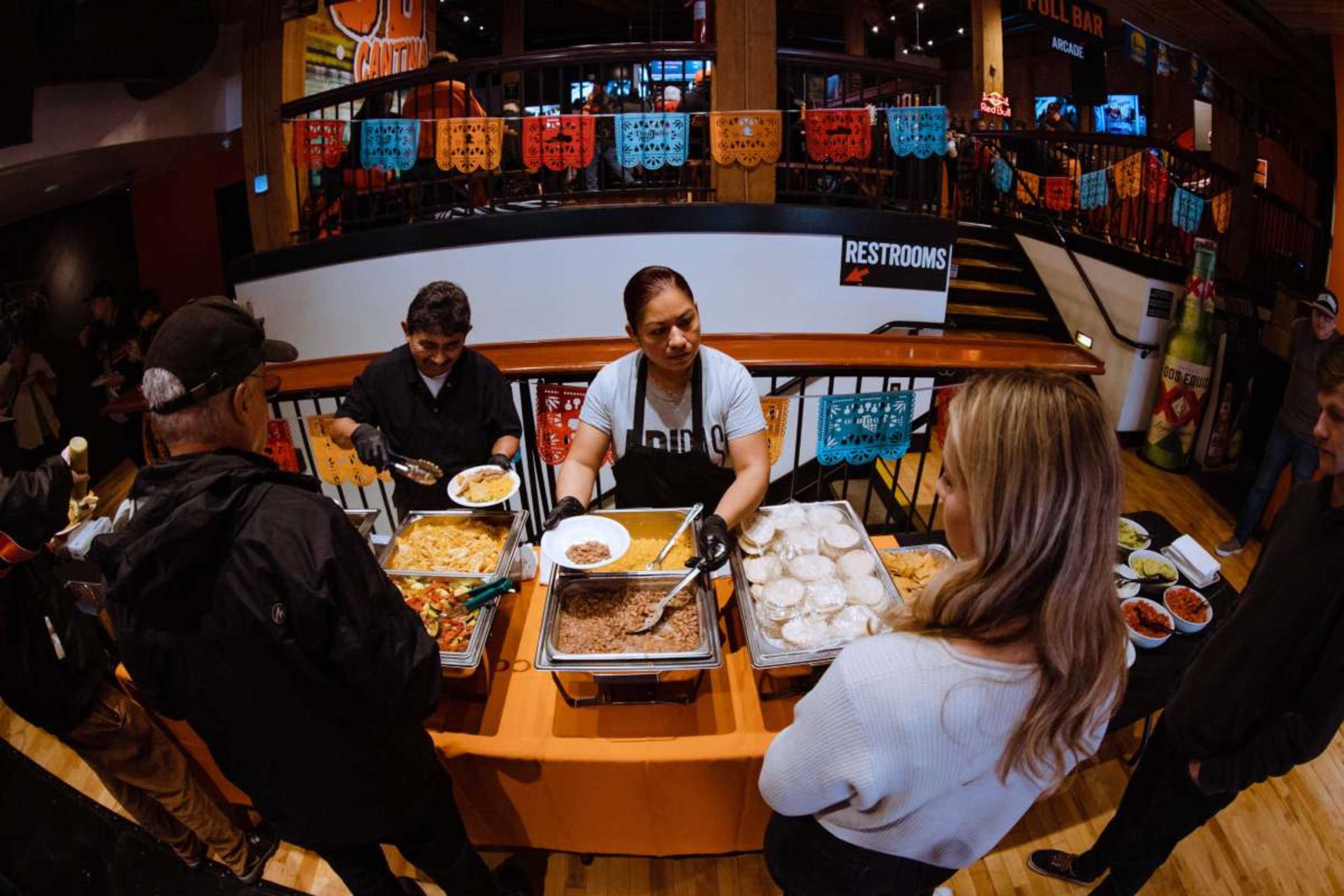 A woman serves food from a counter to customers in a vibrant dining area decorated with colorful papel picado banners.