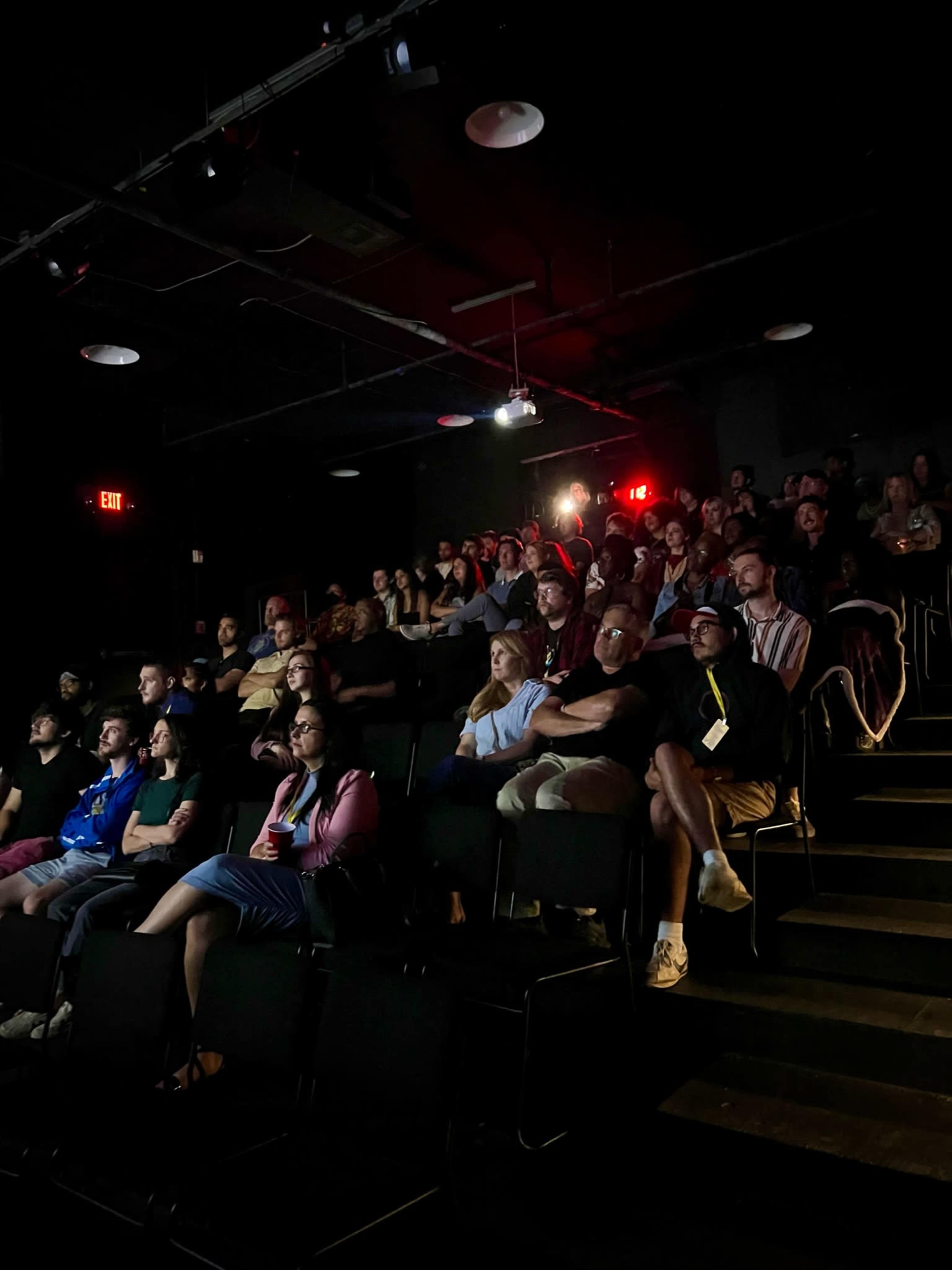 A diverse audience watches a presentation in a dimly lit theater.