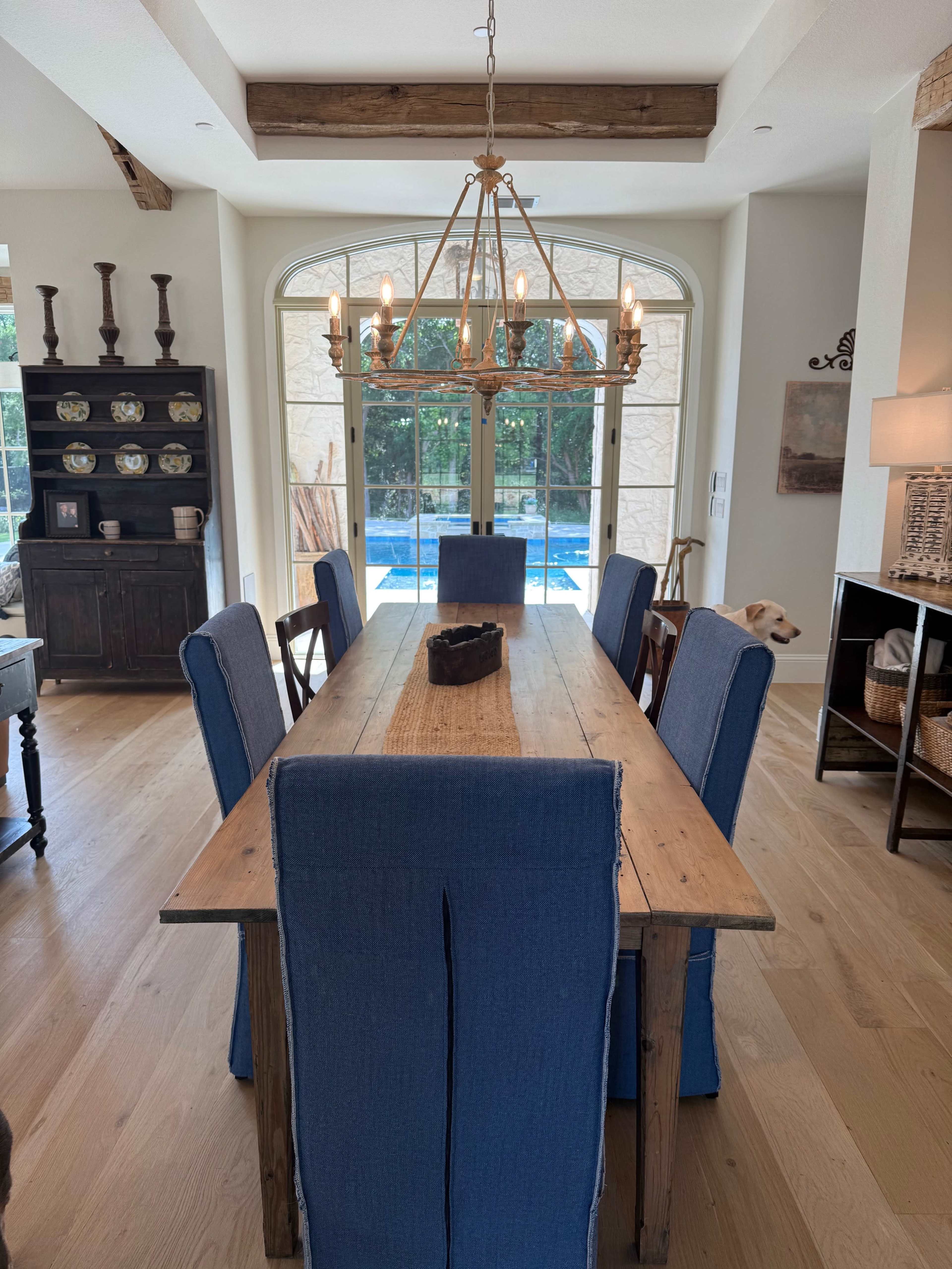 A rustic dining table with blue upholstered chairs is positioned near large windows overlooking a pool area.
