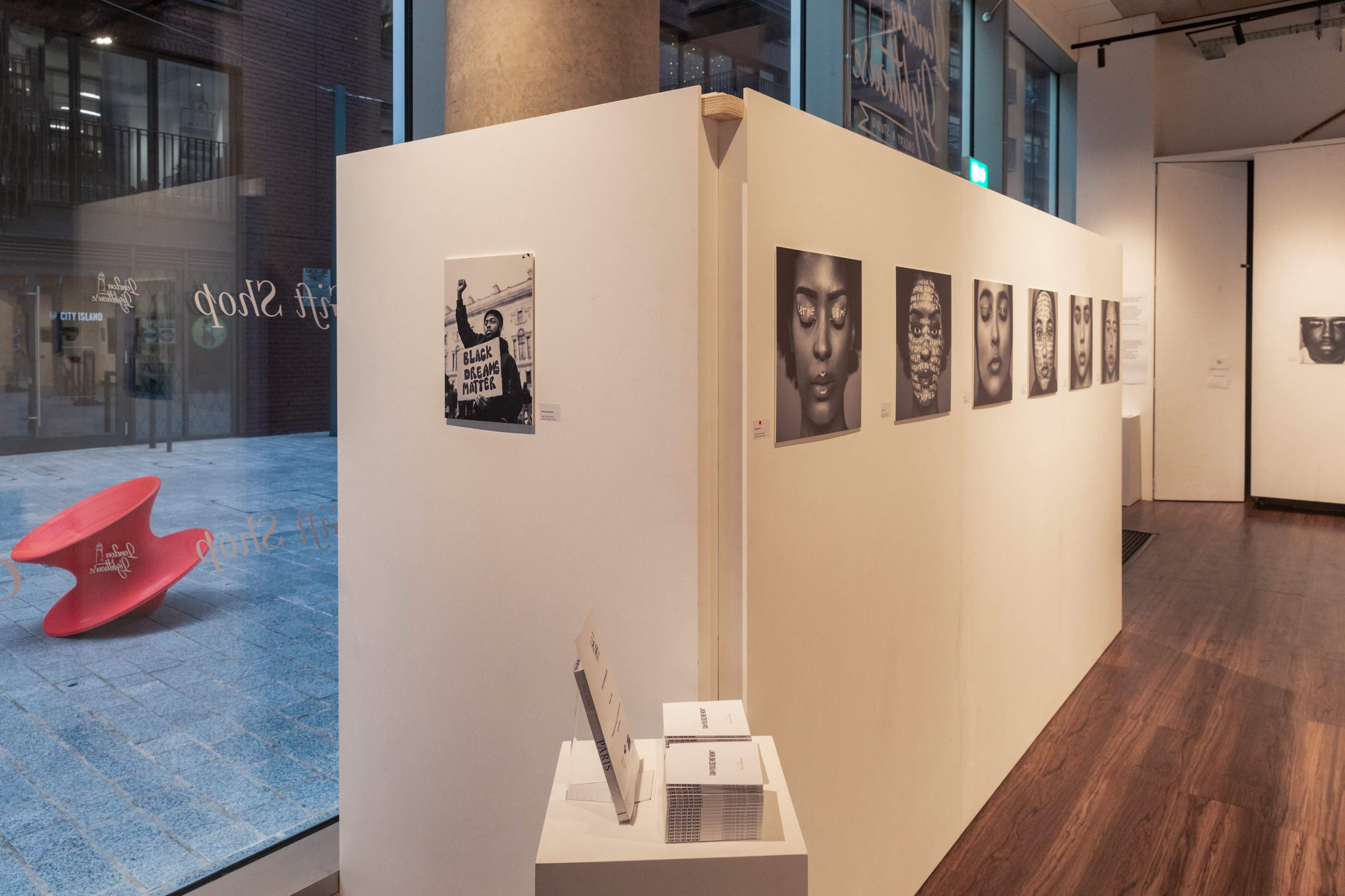 An art gallery interior featuring a white wall displaying black and white photographs, with a pamphlet holder in the foreground.