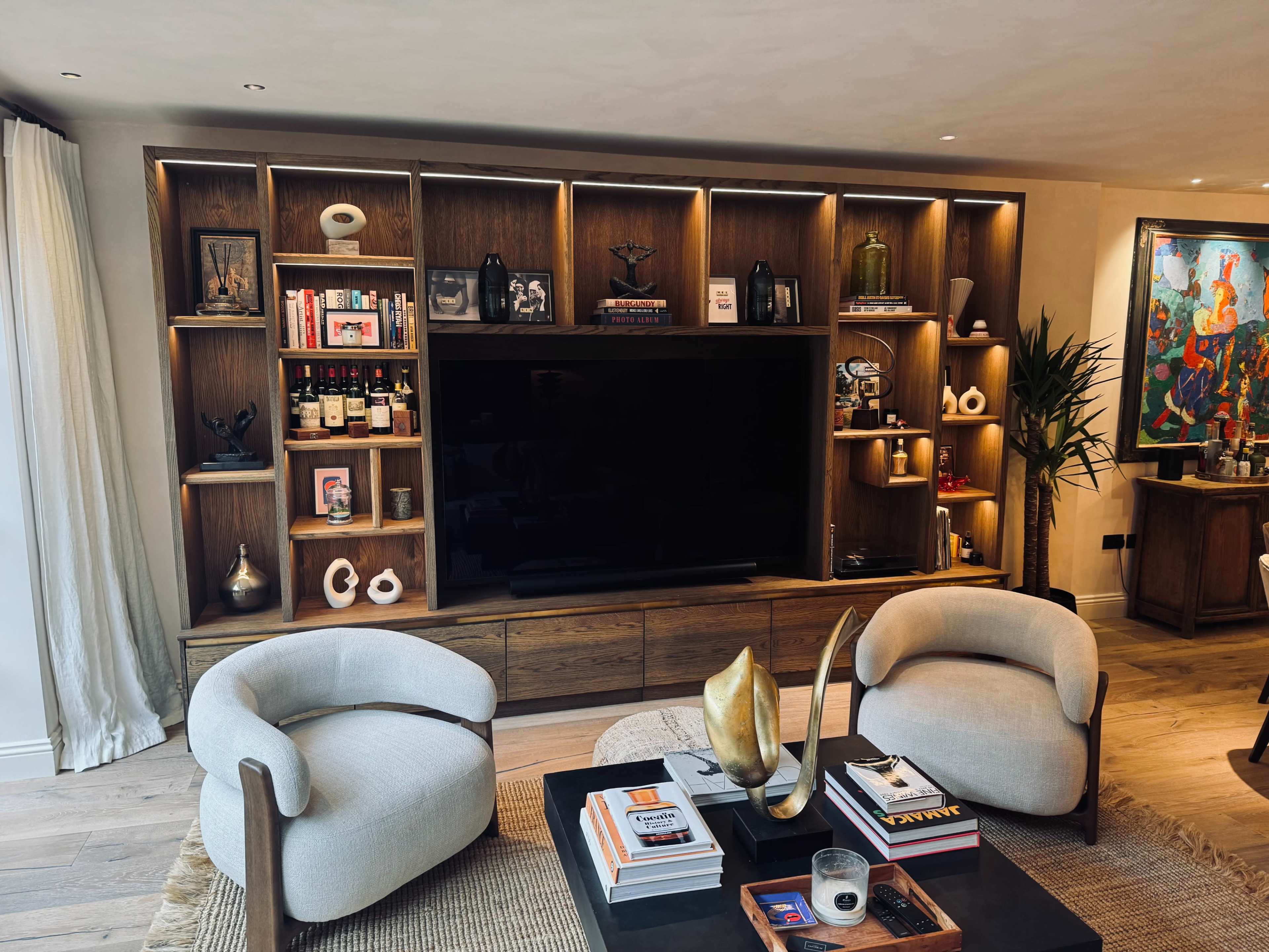 The image shows a modern living room featuring a wooden media unit with illuminated shelves, two light-colored chairs, and a coffee table adorned with books and decorative items.