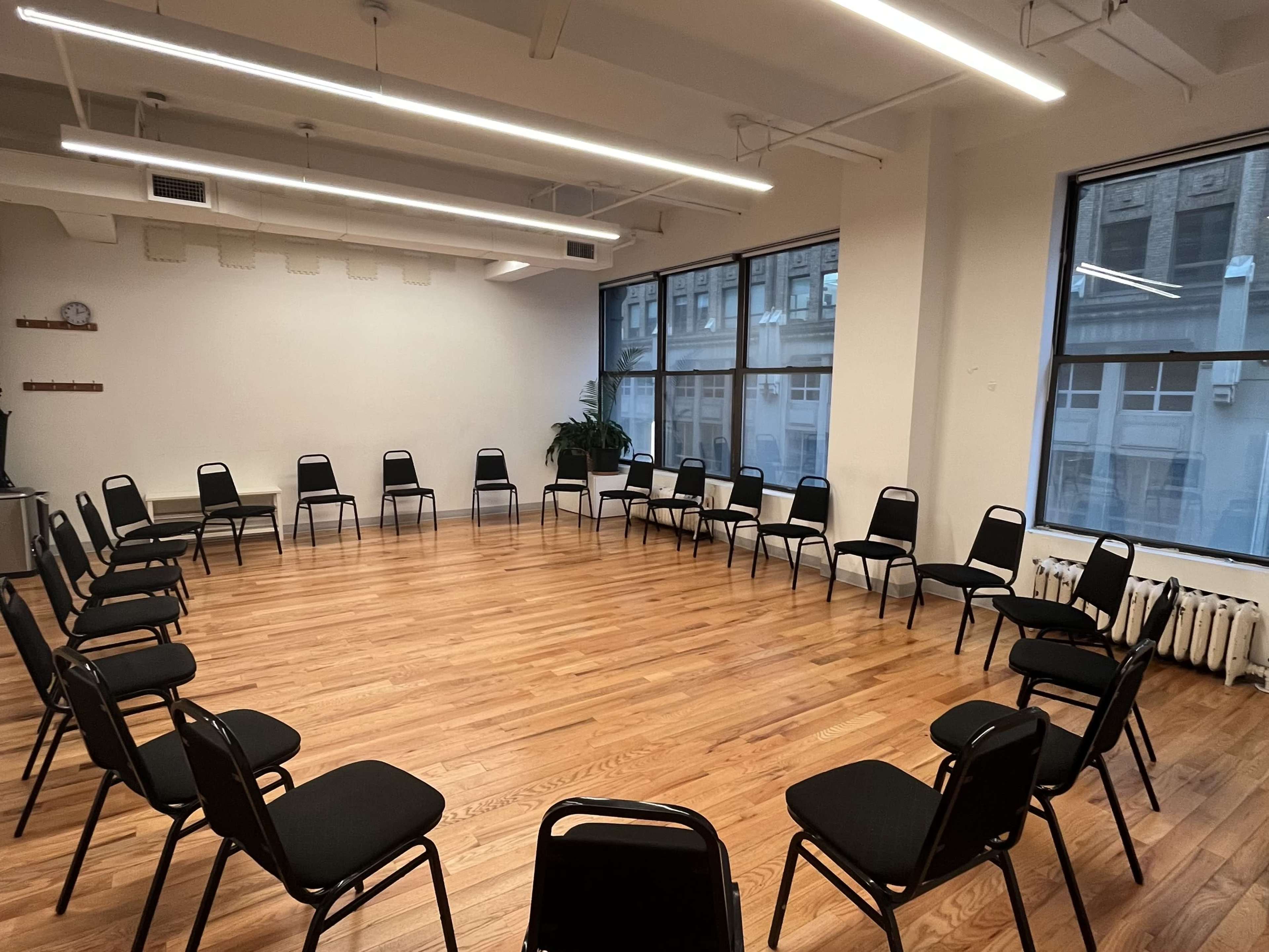 A spacious room with wooden floors contains two circular arrangements of black chairs facing each other, surrounded by large windows.