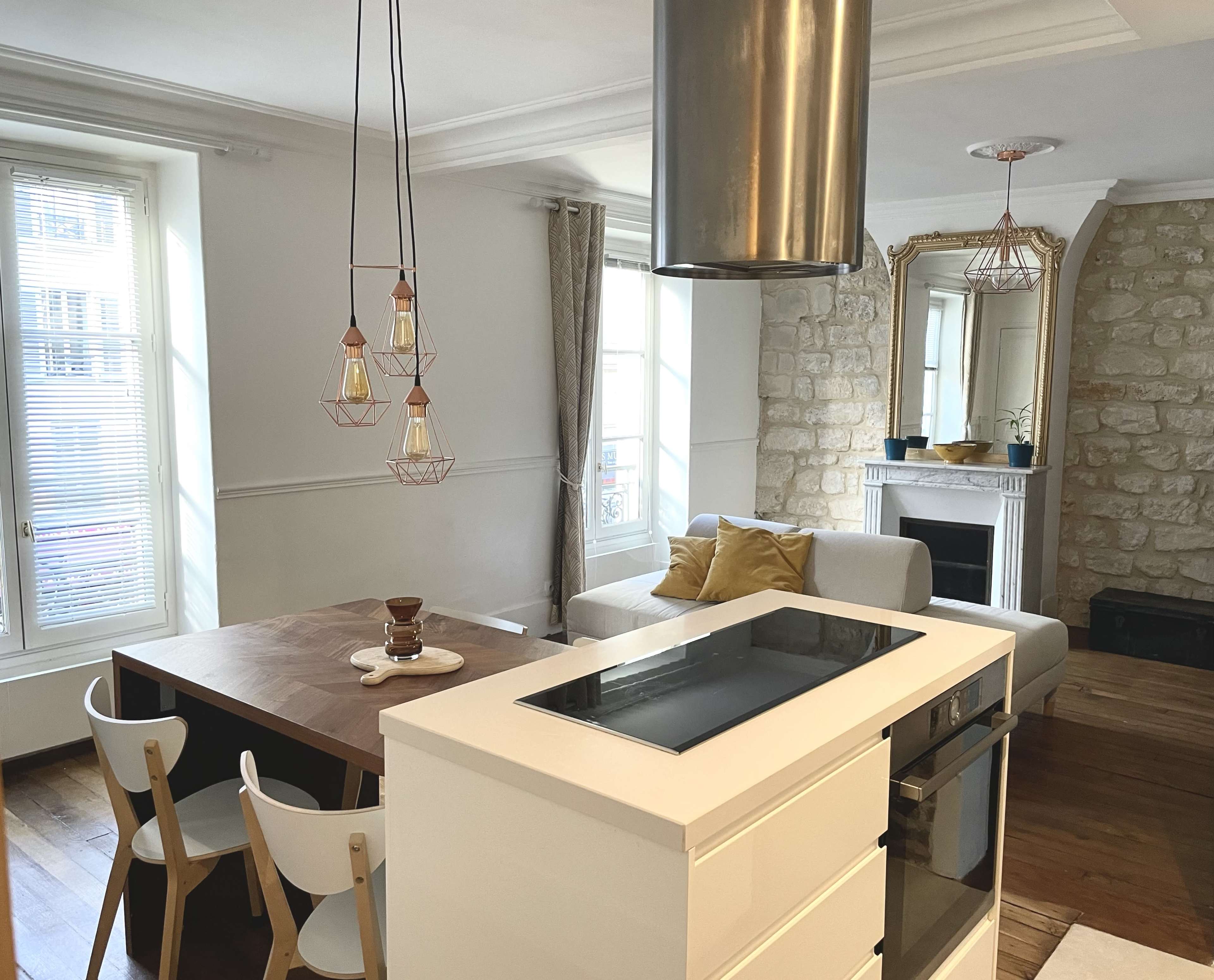The image shows a modern kitchen and living area with a wooden dining table, a white couch, and large windows offering natural light.
