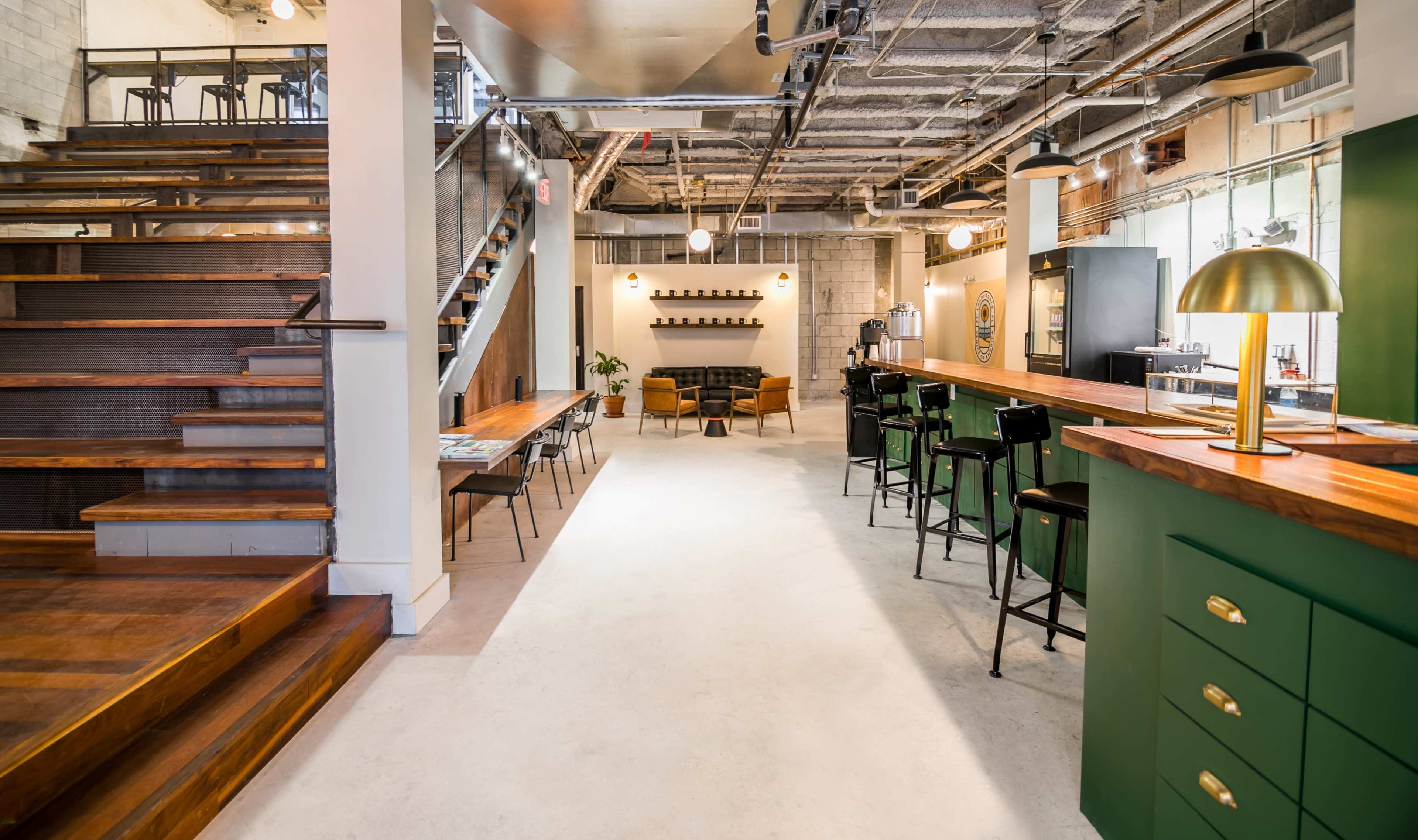 A modern interior space featuring a staircase, a wooden bar with green accents, seating areas, and a minimalist design.
