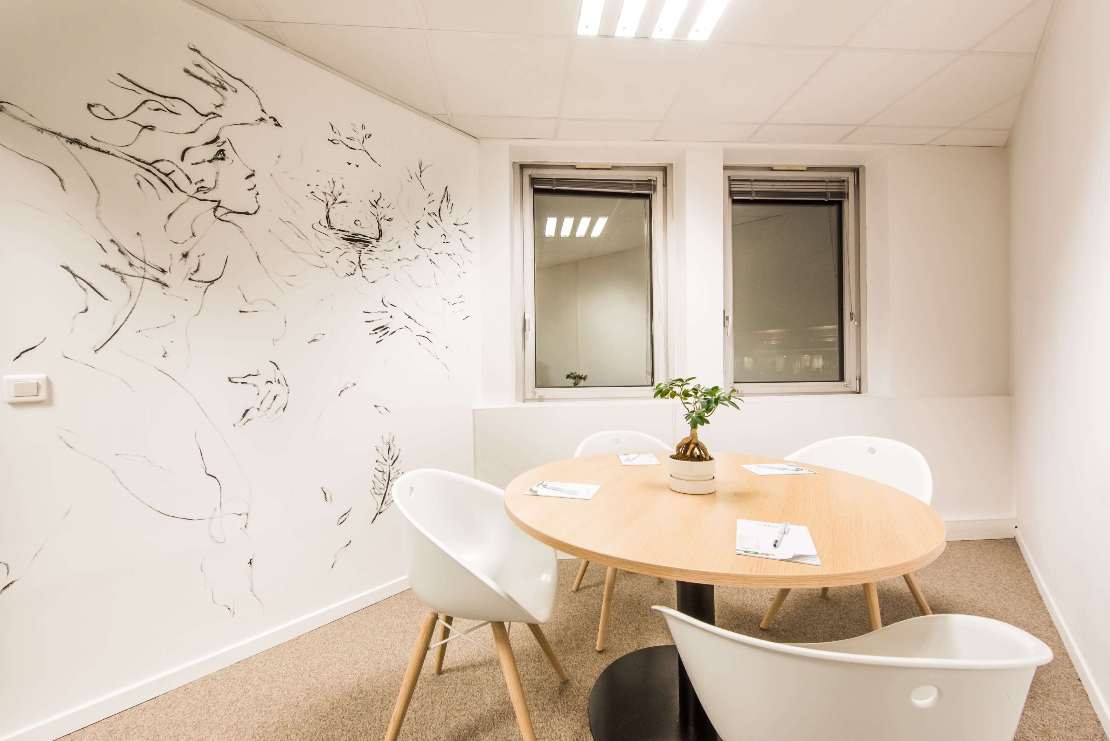 A small meeting room features a round wooden table surrounded by white chairs, with a plant at the center and a mural on the wall behind it.