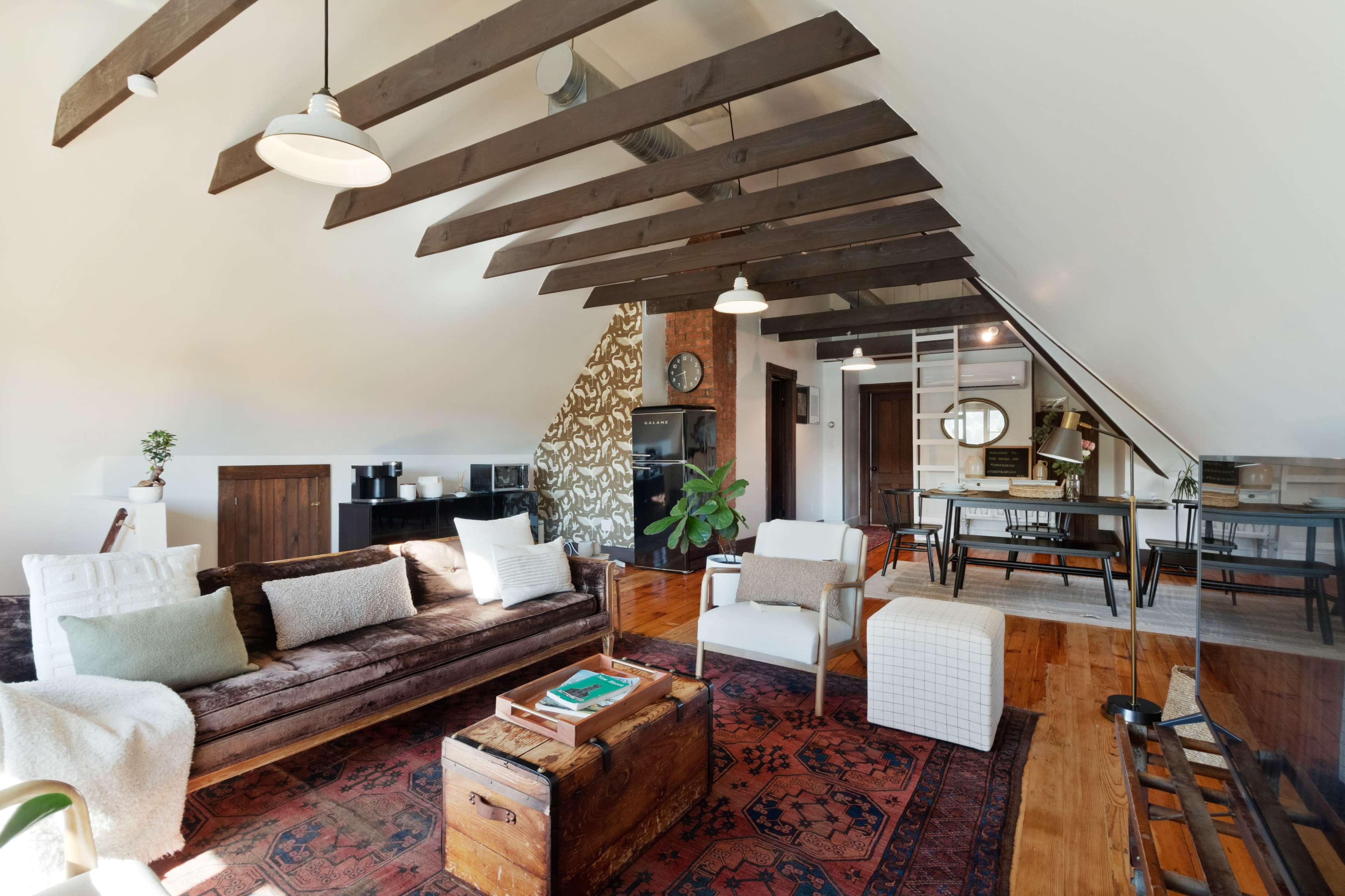 A cozy loft space features a brown sofa, wooden beams on the ceiling, a coffee table, and a dining area with a black fridge in the background.