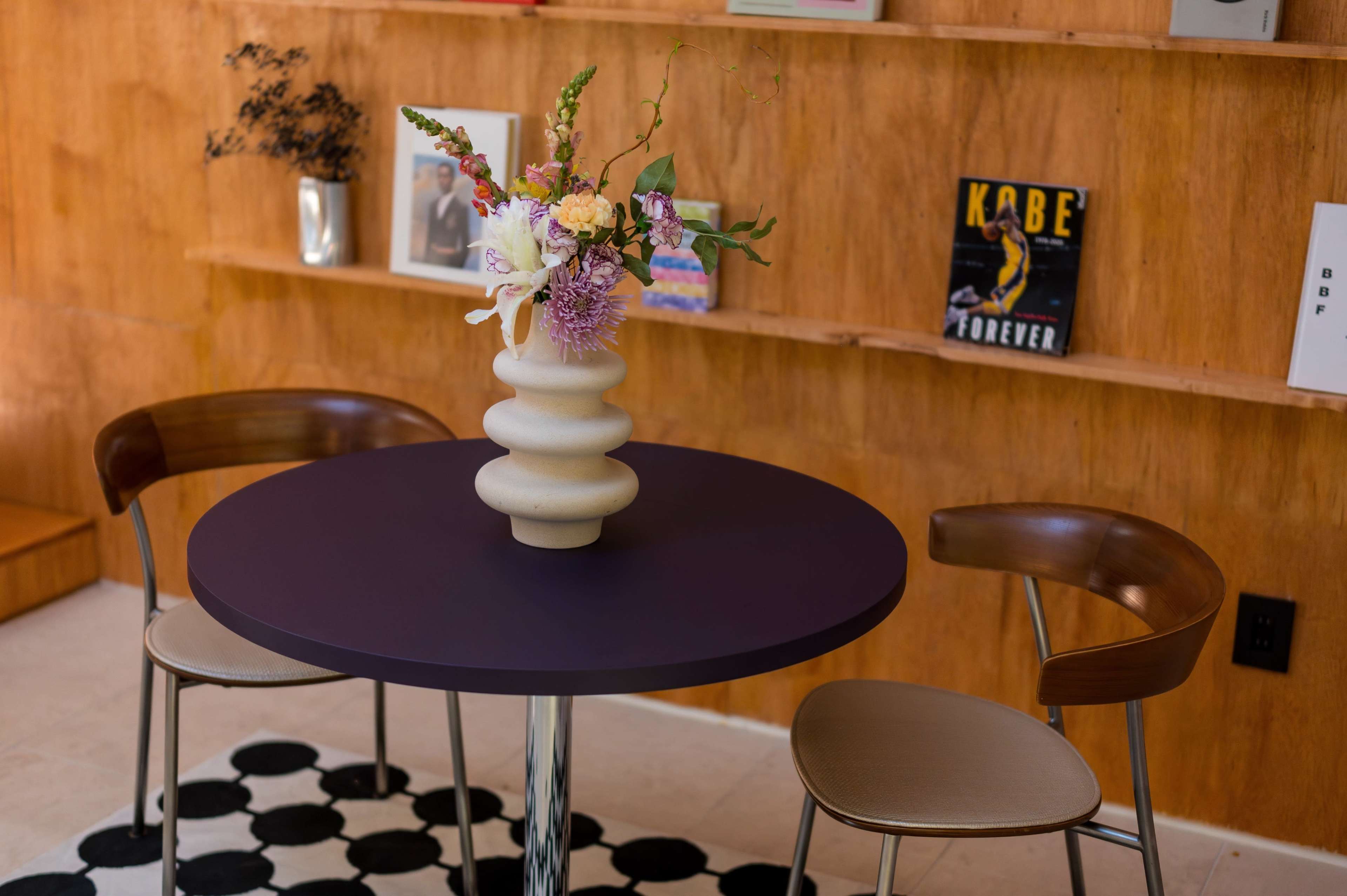 A round purple table with a decorative vase of flowers sits between two wooden chairs in a room with a wooden wall and shelves displaying various items.