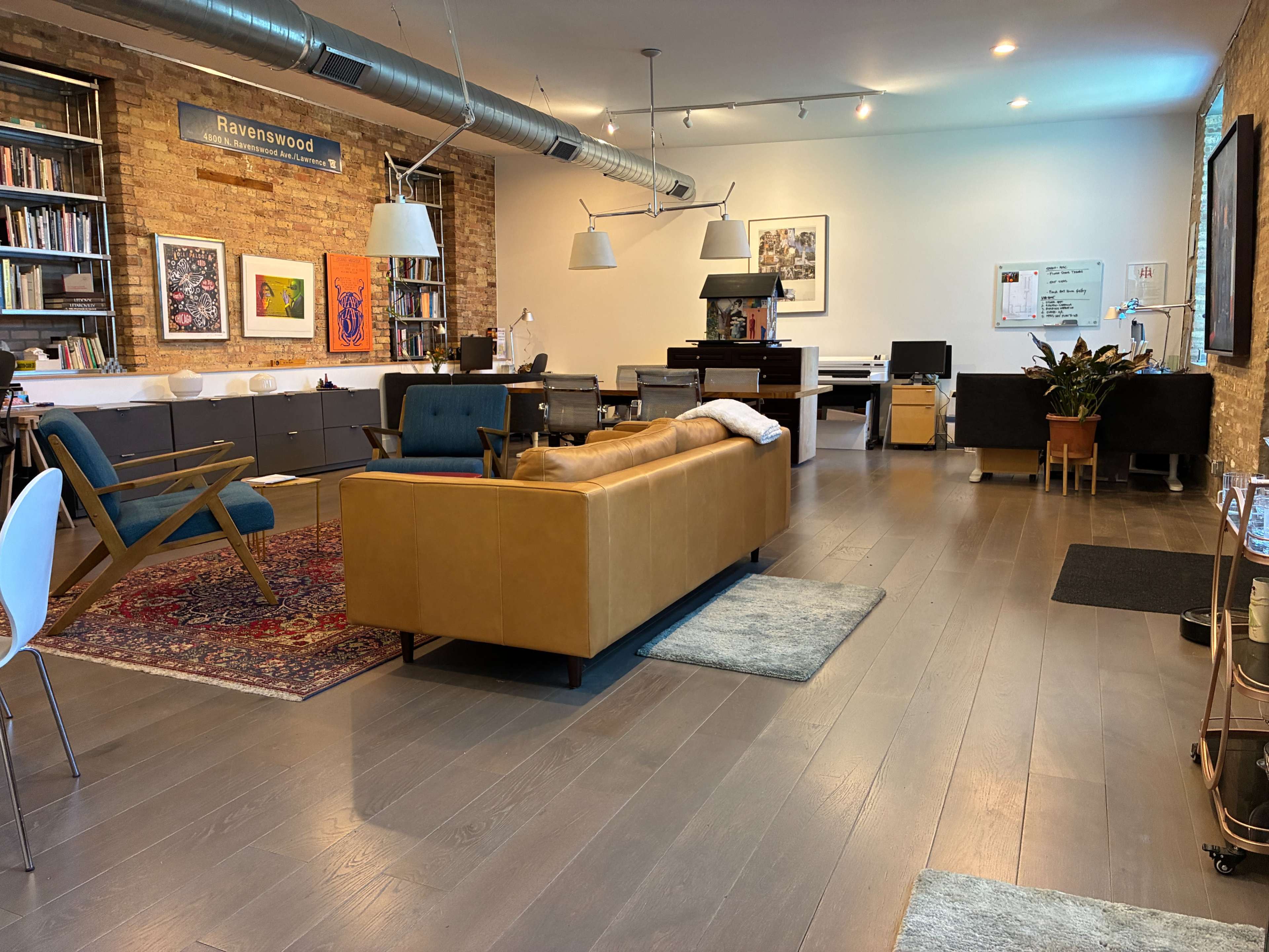 The image shows a spacious loft interior featuring a brown leather sofa, two blue chairs, a large bookshelf, and a workspace with a computer and desk.