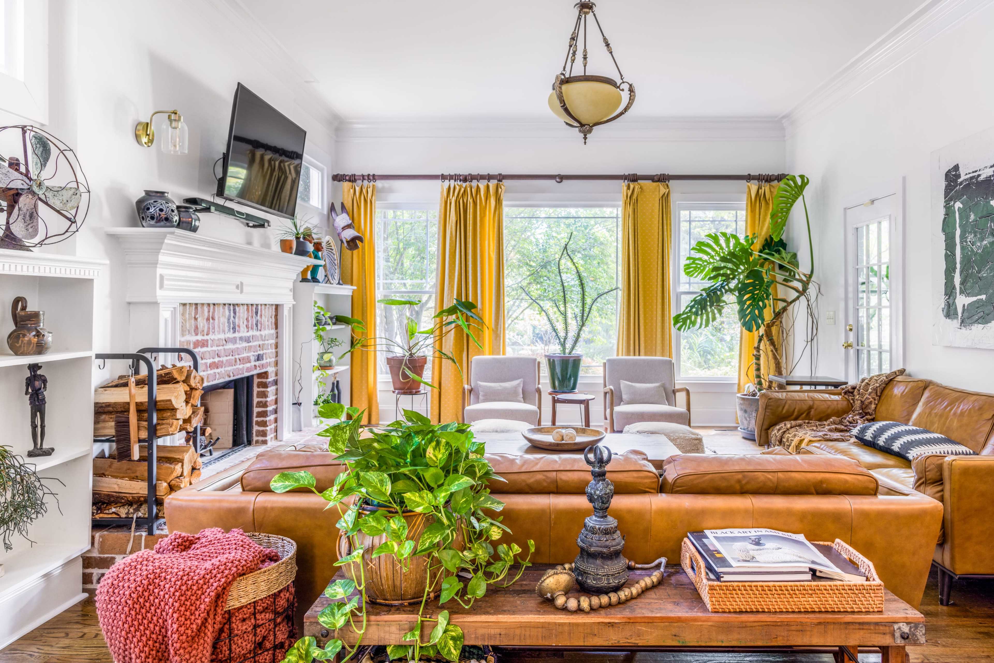 A bright living room features a brown leather sofa, a wooden coffee table, and large windows adorned with yellow curtains, surrounded by indoor plants.