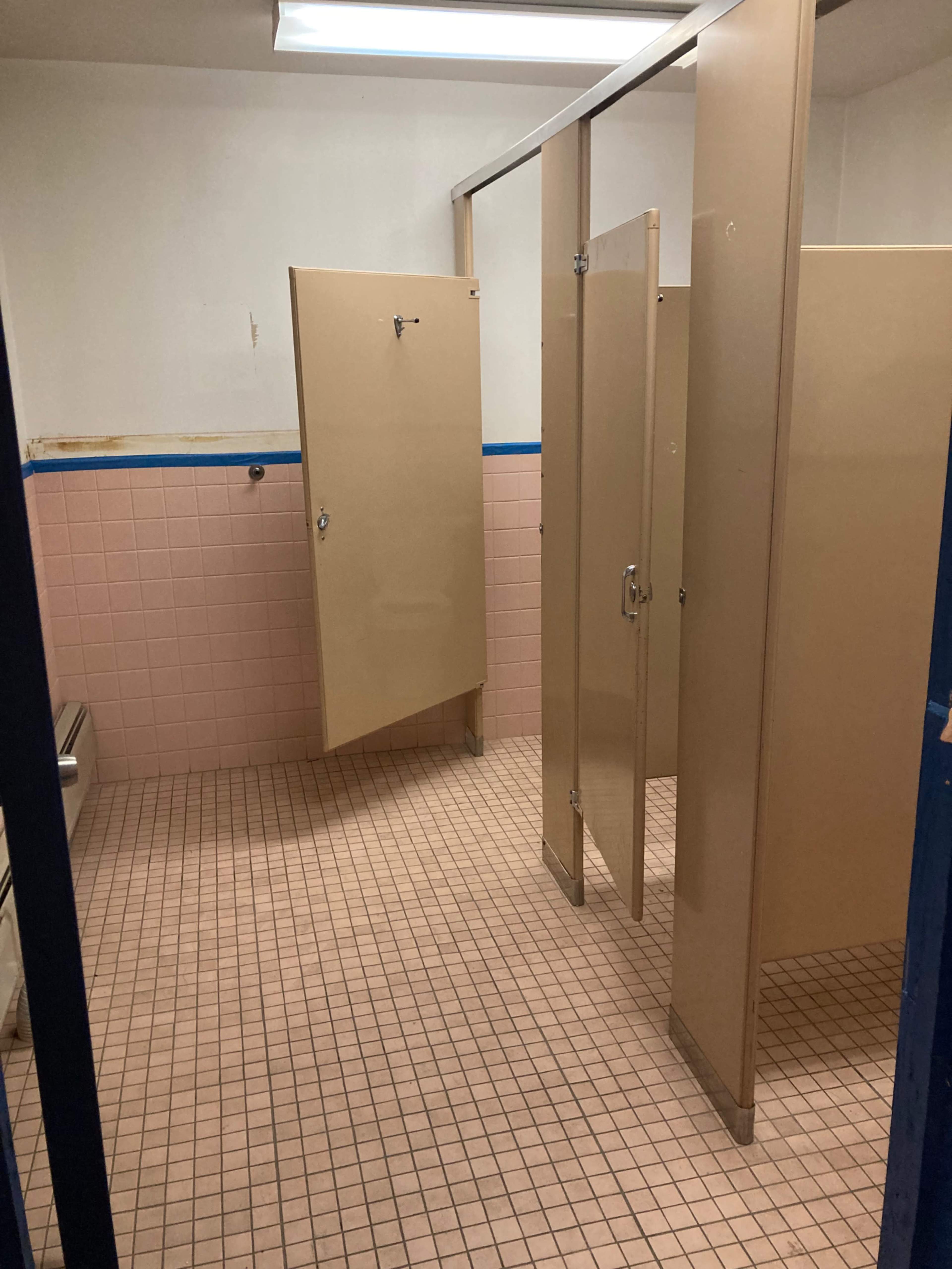 A restroom with light-colored tiled flooring and several beige stall doors partially closed.