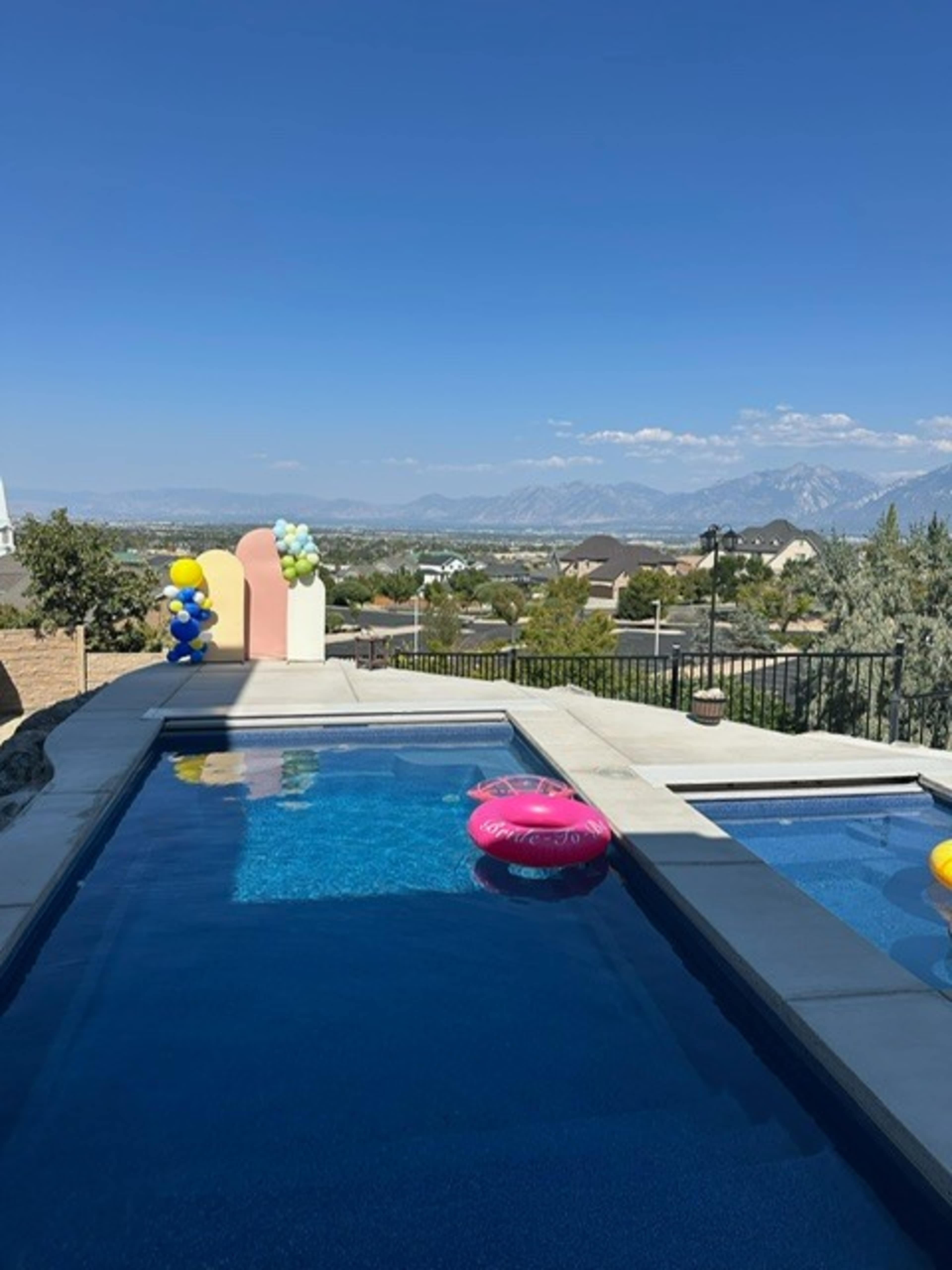 A clear swimming pool with colorful floats overlooks a scenic view of mountains and homes under a bright blue sky.