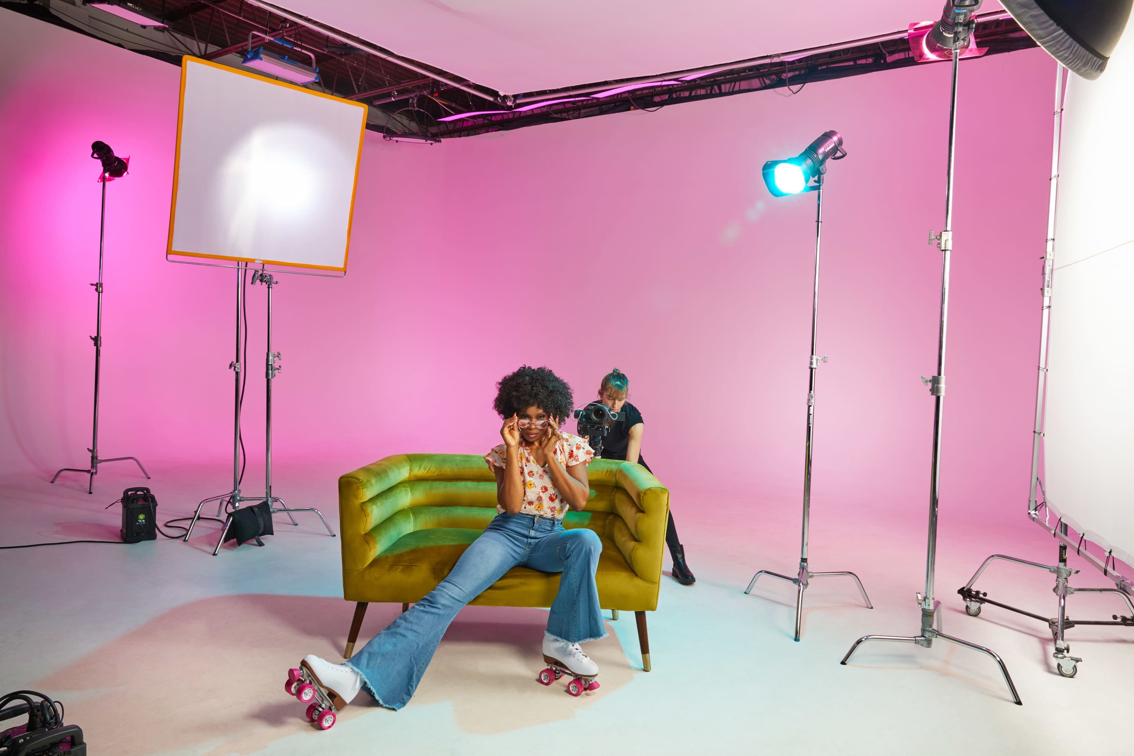 A person wearing roller skates sits on a green couch in a studio with pink walls and multiple lights set up for a photoshoot.