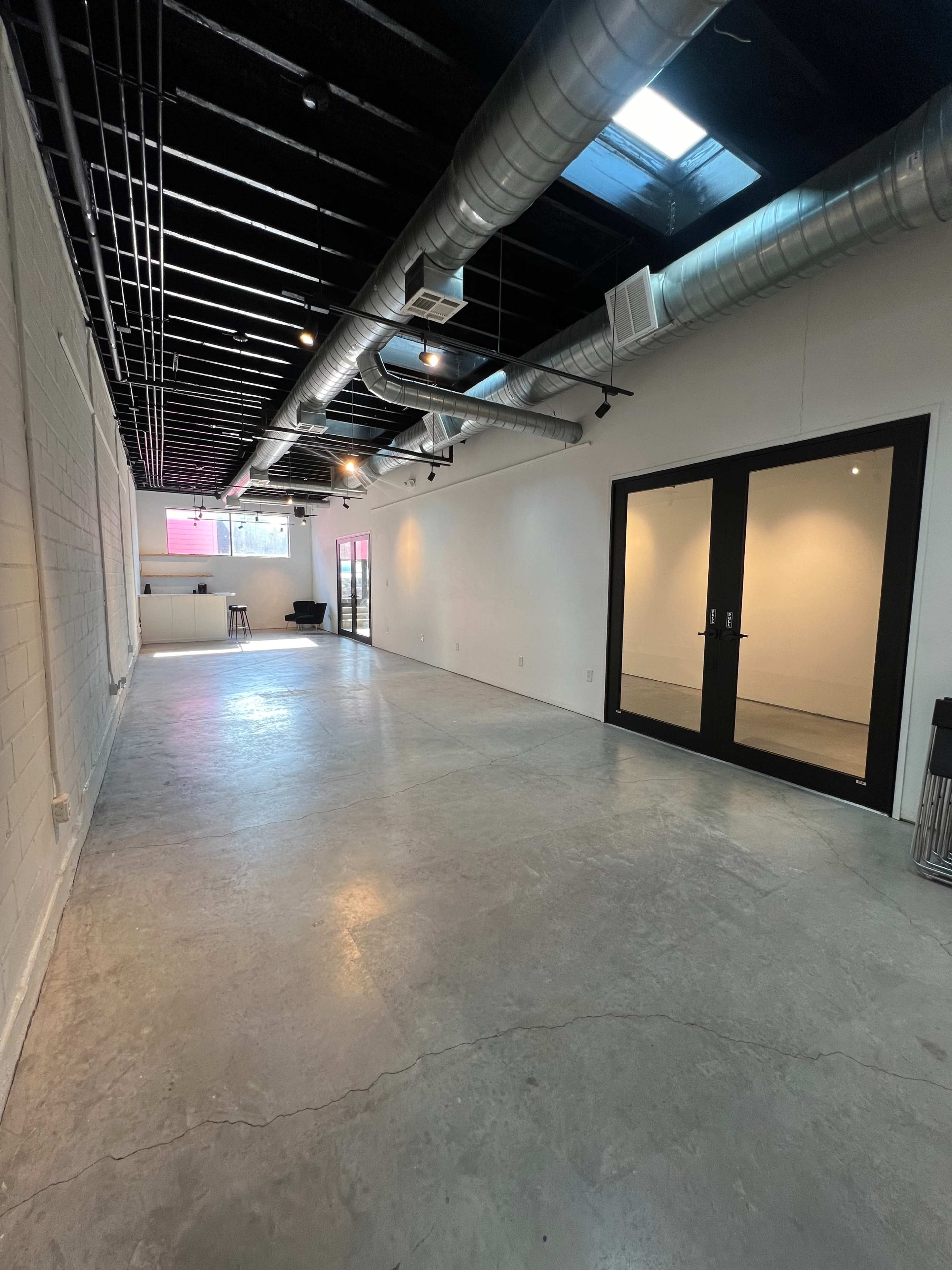 The image shows a spacious, empty interior room with concrete flooring, exposed ductwork, and large glass doors.