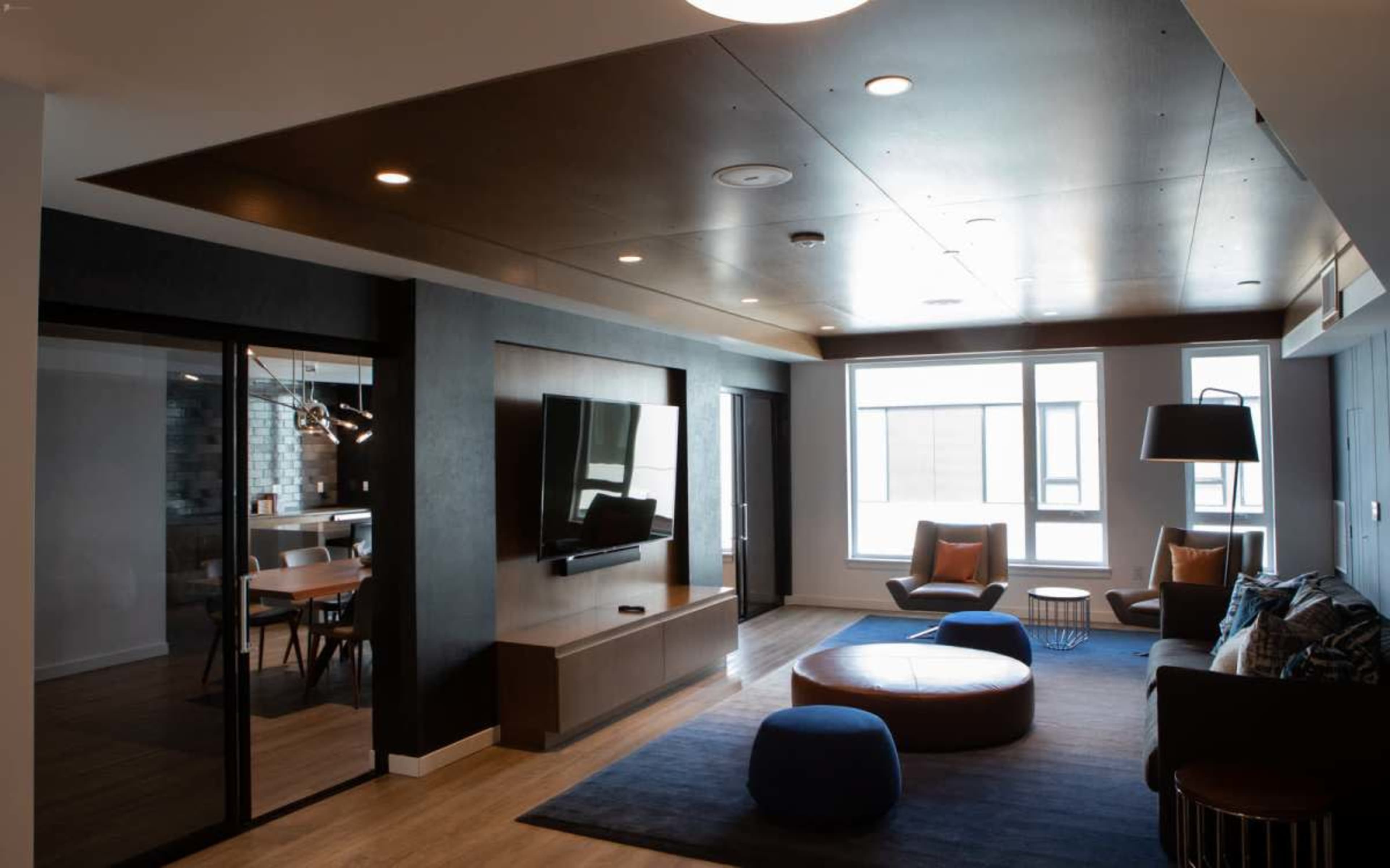 The image shows a modern living space featuring a lounge area with seating, a television mounted on the wall, and large windows allowing natural light.