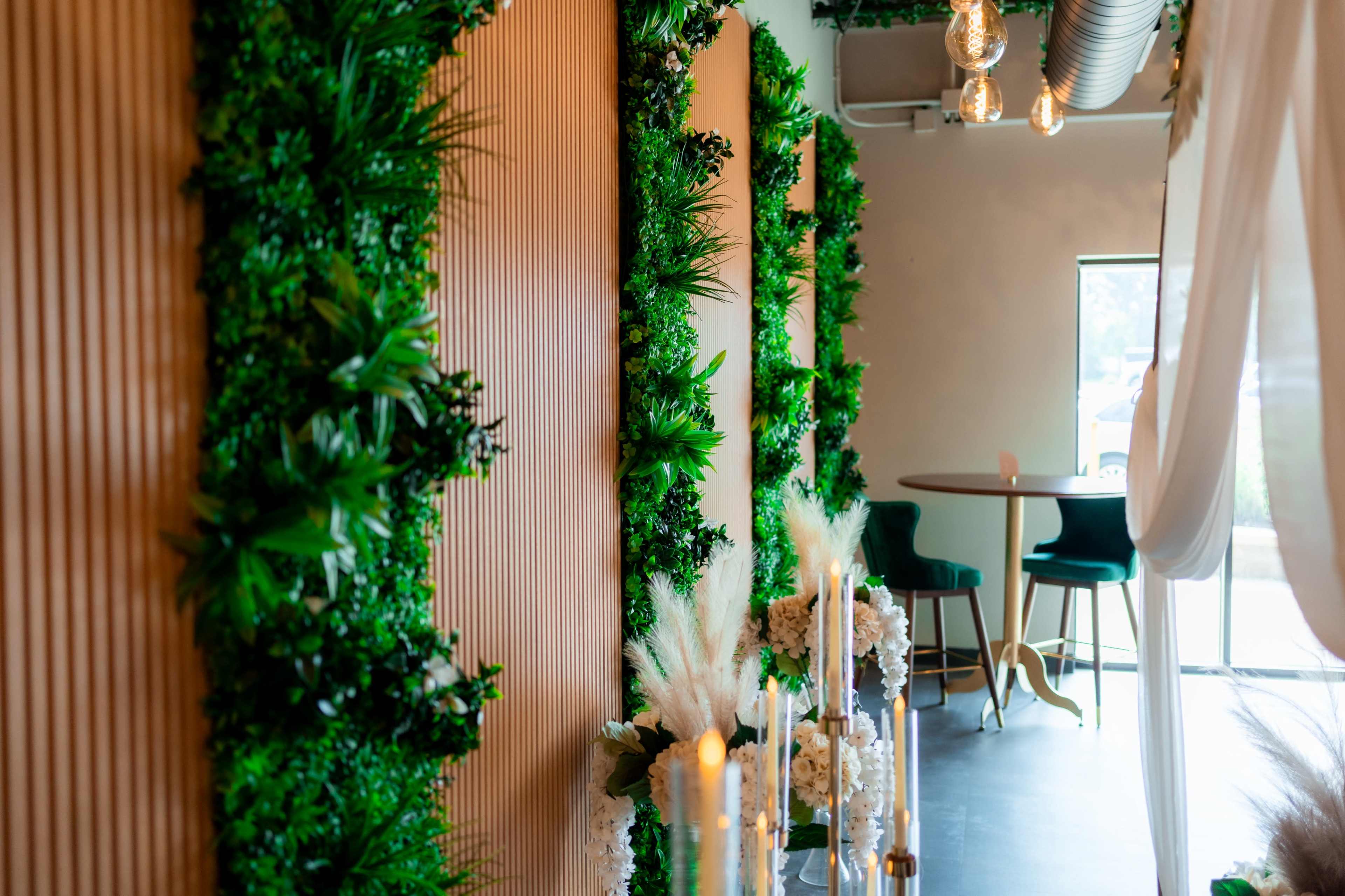 The image shows a decorated interior space featuring green plants on wood-paneled walls, a small round table with chairs in the background, and candles arranged in front of a sheer curtain.